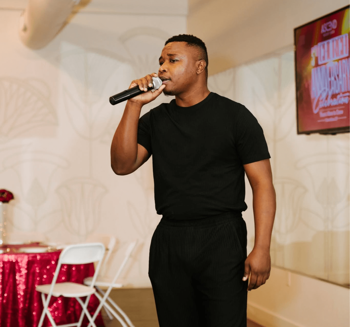 Man holding microphone, speaking or singing, wearing black shirt and pants, in indoor setting with decorated wall and TV screen