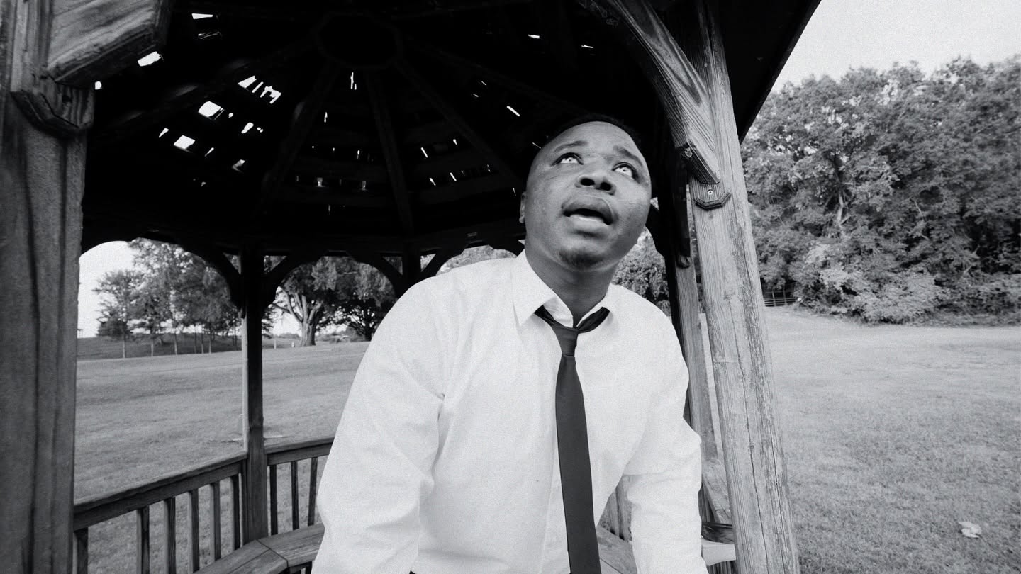 Person with surprised expression standing under a wooden gazebo outdoors, wearing a white shirt and tie, trees in background.