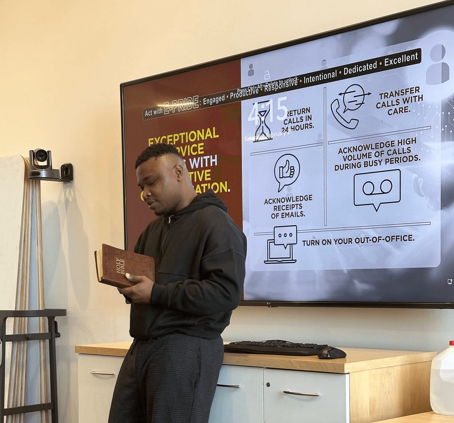 Person standing in front of a presentation screen, holding a book, with a cabinet and a water bottle nearby.