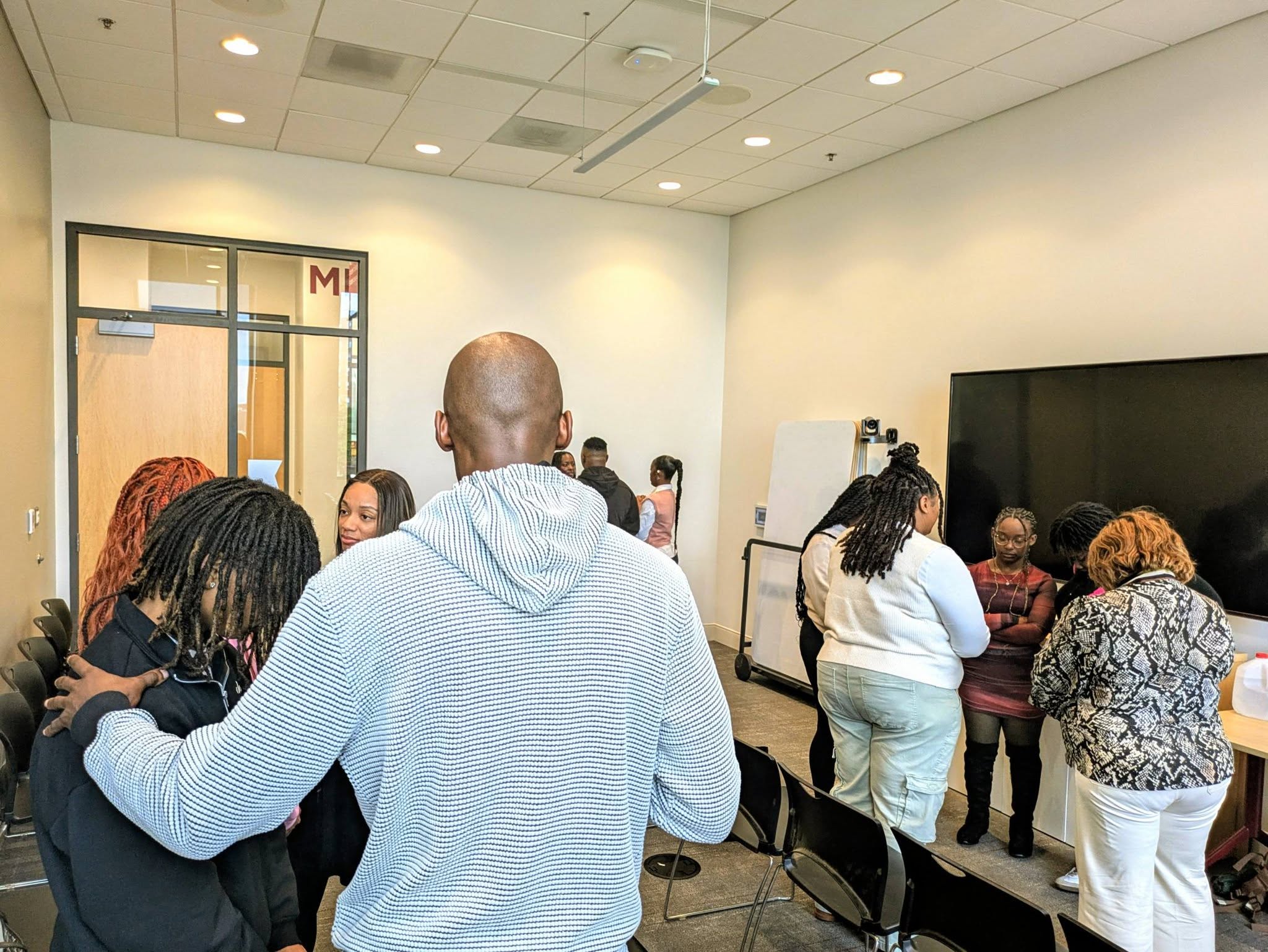 Group of people gathered in a room with white walls and a large screen, some talking and others listening.
