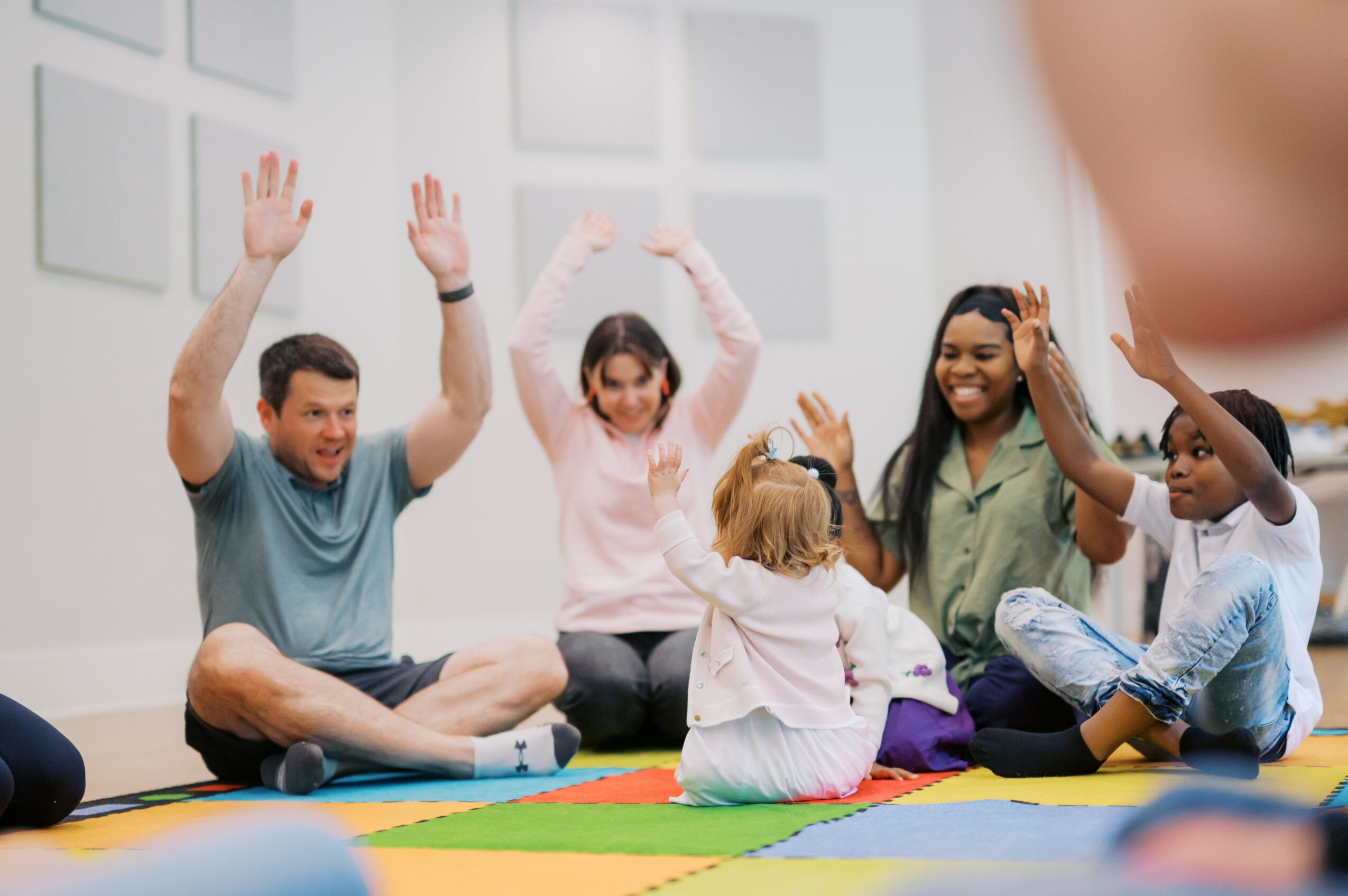 Group of five people sitting on colorful mats, raising hands, in a bright room with white walls.