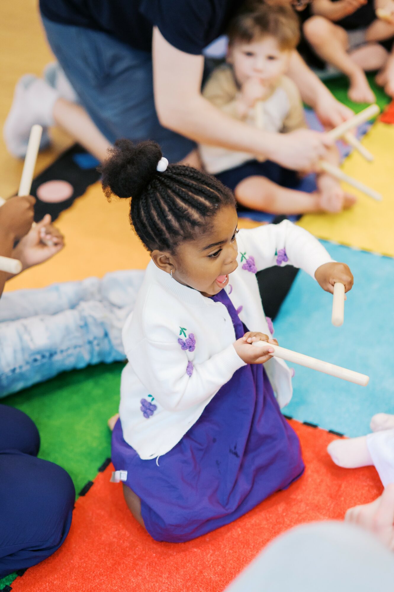 Young girl with braided hair sitting on colorful mat, holding a white stick, surrounded by children and adults, engaging in activity.