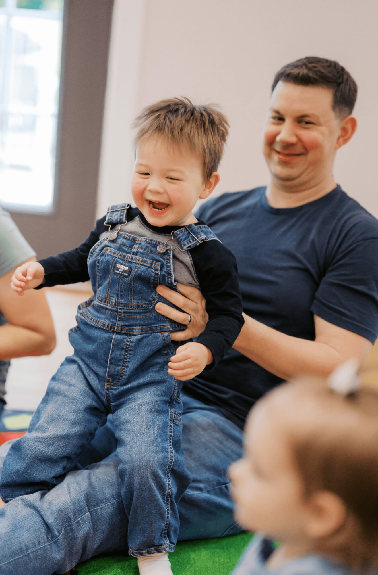 Adult holding smiling child in denim overalls indoors, with other children nearby, bright window in background.