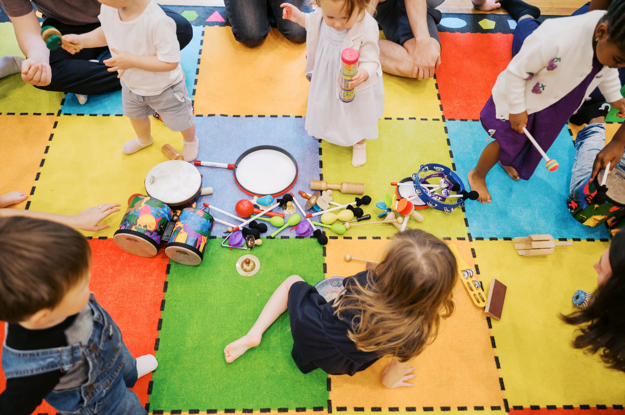 Children sitting on colorful mats with drums, toys, and a girl holding a bottle, seen from above.