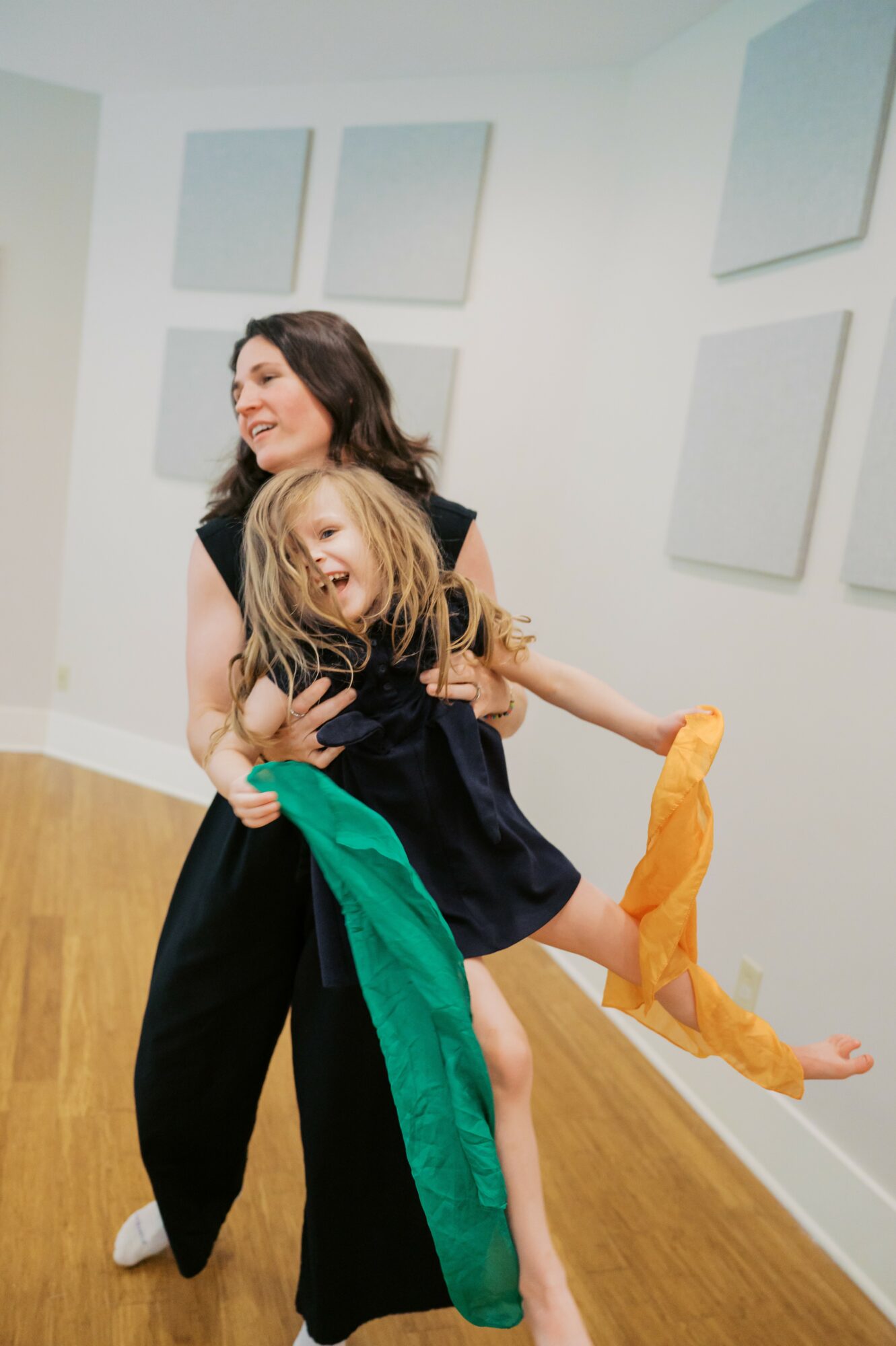 Two people, a woman and a girl, smiling and hugging in a room with white walls and wooden floor.