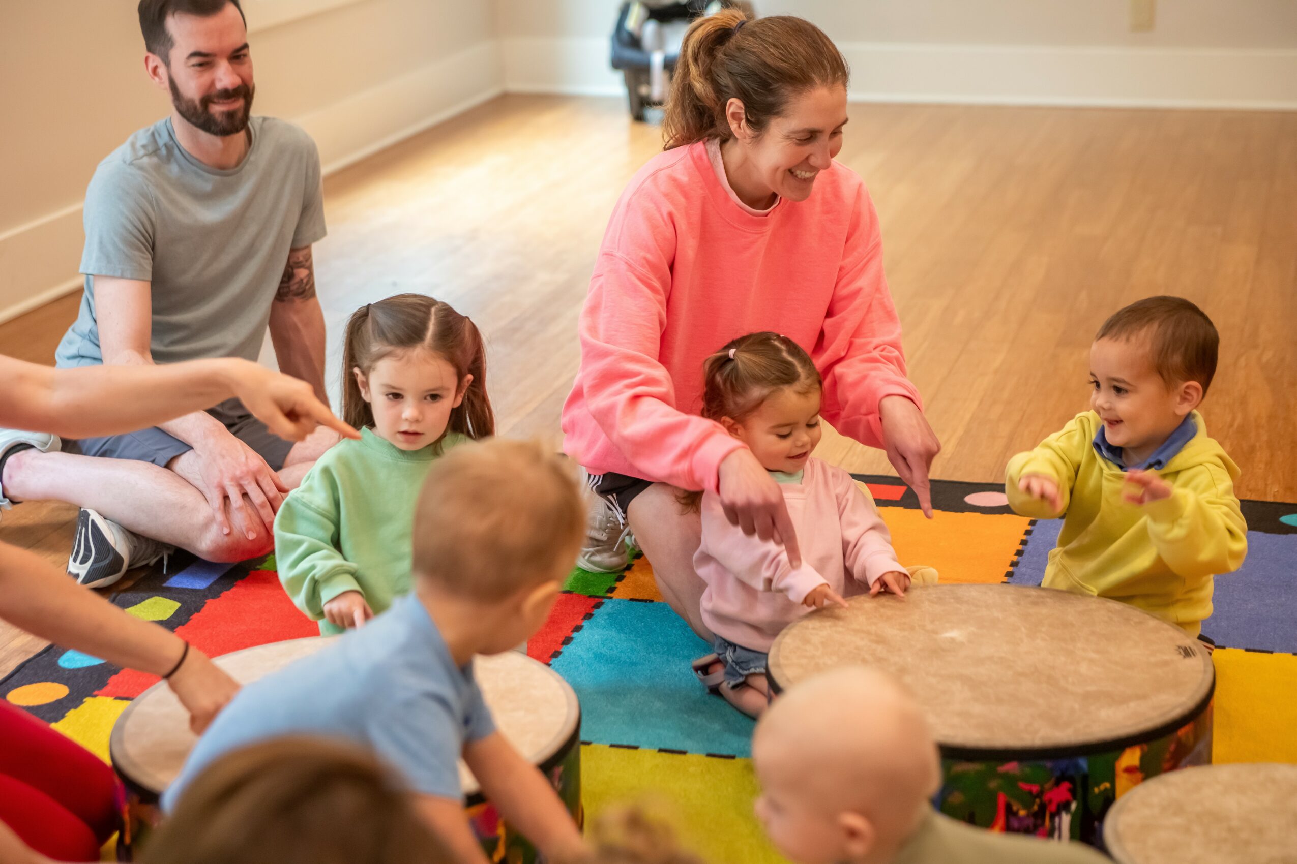 Group of children and two adults sitting on a colorful mat in a room, engaging in a group activity around a small table.