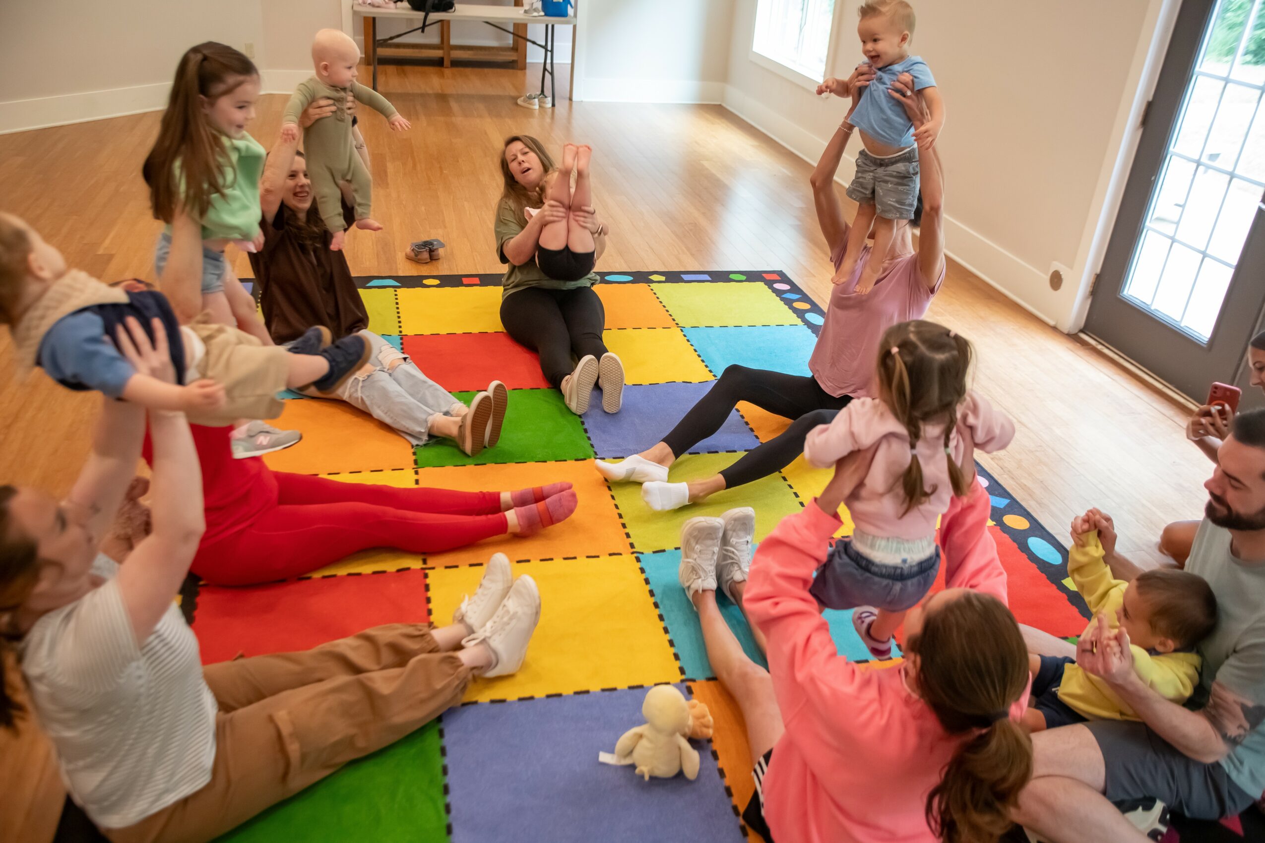 Children and adults sit on colorful foam mats in a room, engaging in a group activity with toys and stuffed animals.