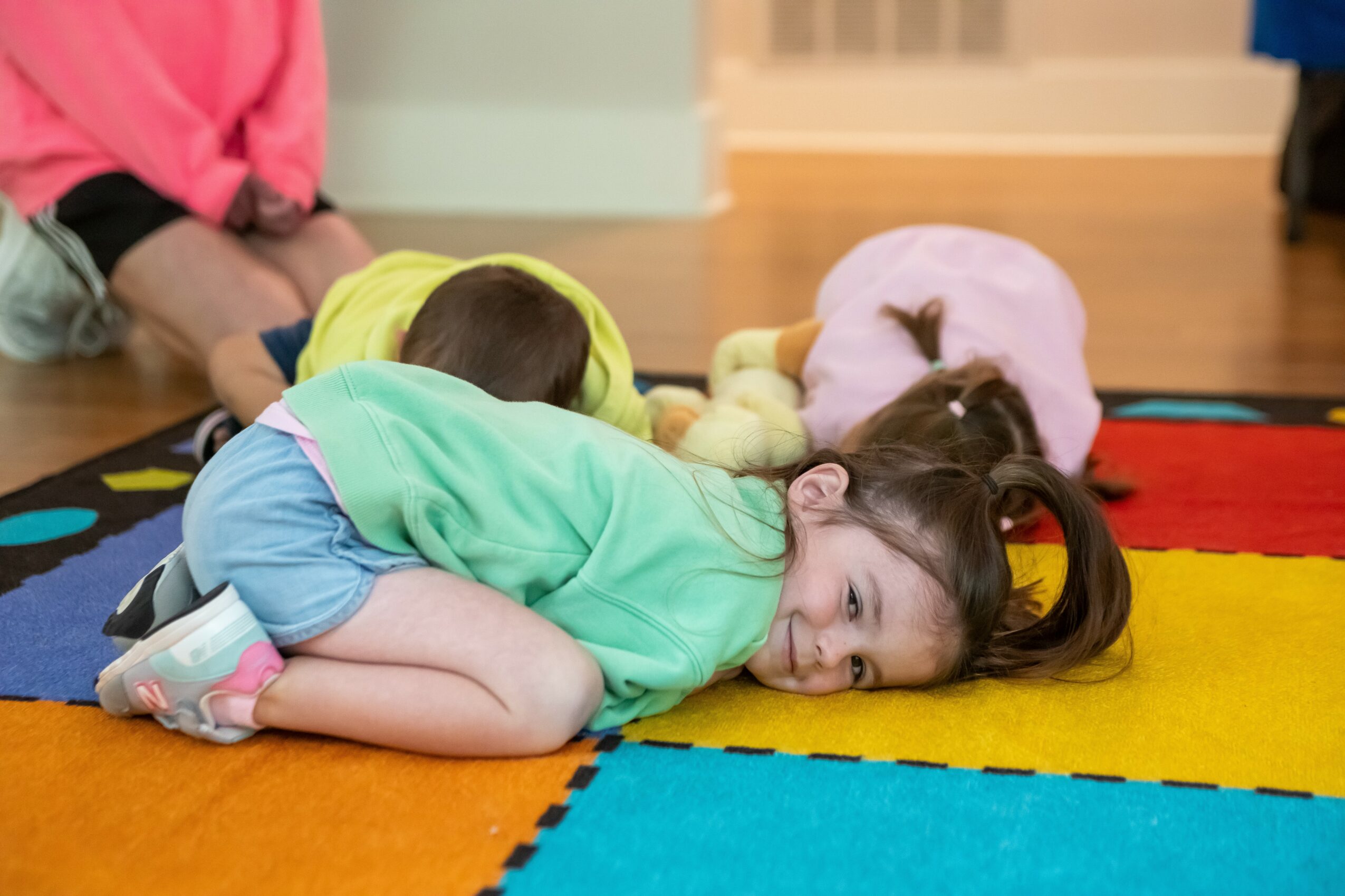 Child lying on colorful carpet, smiling, with other children in the background, indoors.
