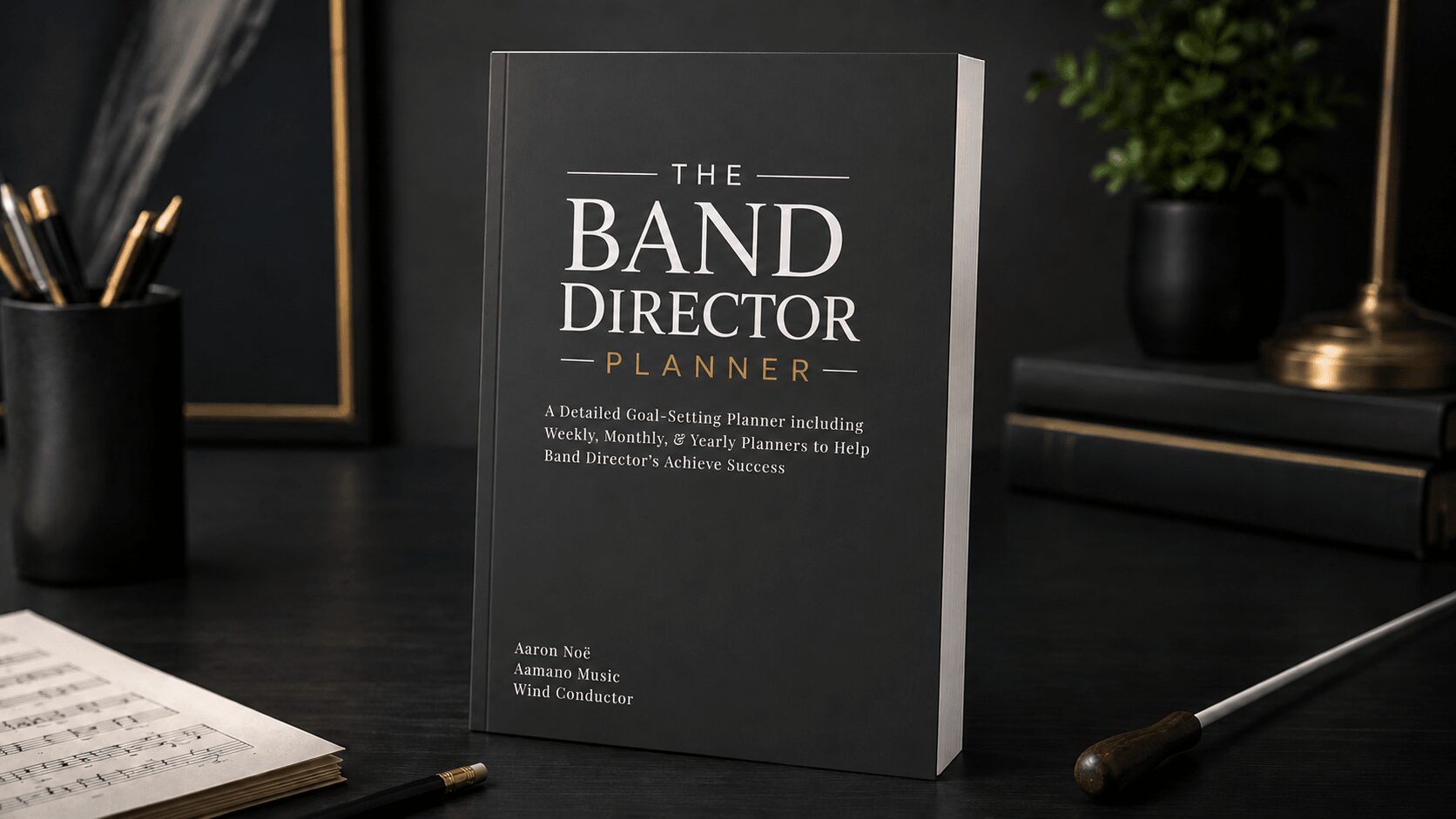 Book titled 'The Band Director Planner' on a desk with pens, notebook, and plant in background.