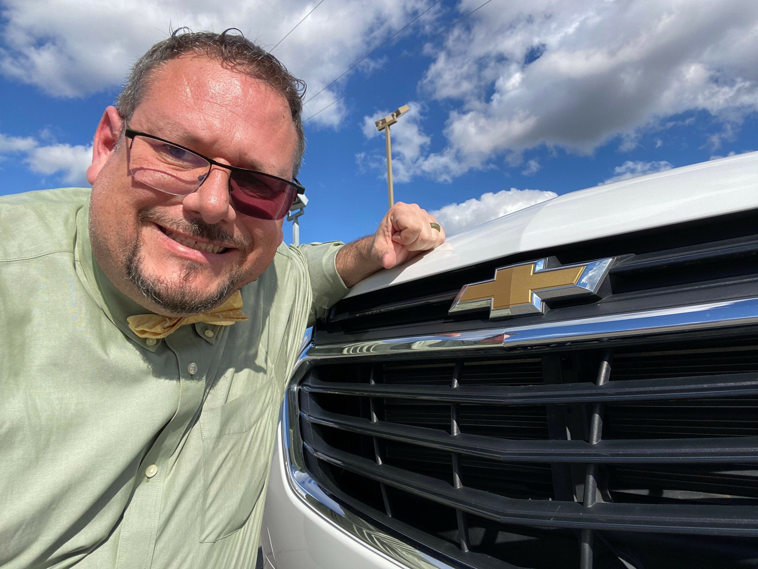 Man with sunglasses and a green shirt stands next to a vehicle with a Chevrolet logo, holding a small flag, against a blue sky with clouds.