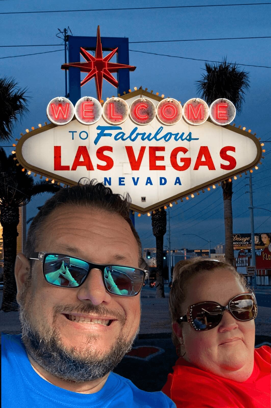 Two people smiling in front of the Las Vegas welcome sign at dusk, with palm trees and a blue sky in background.