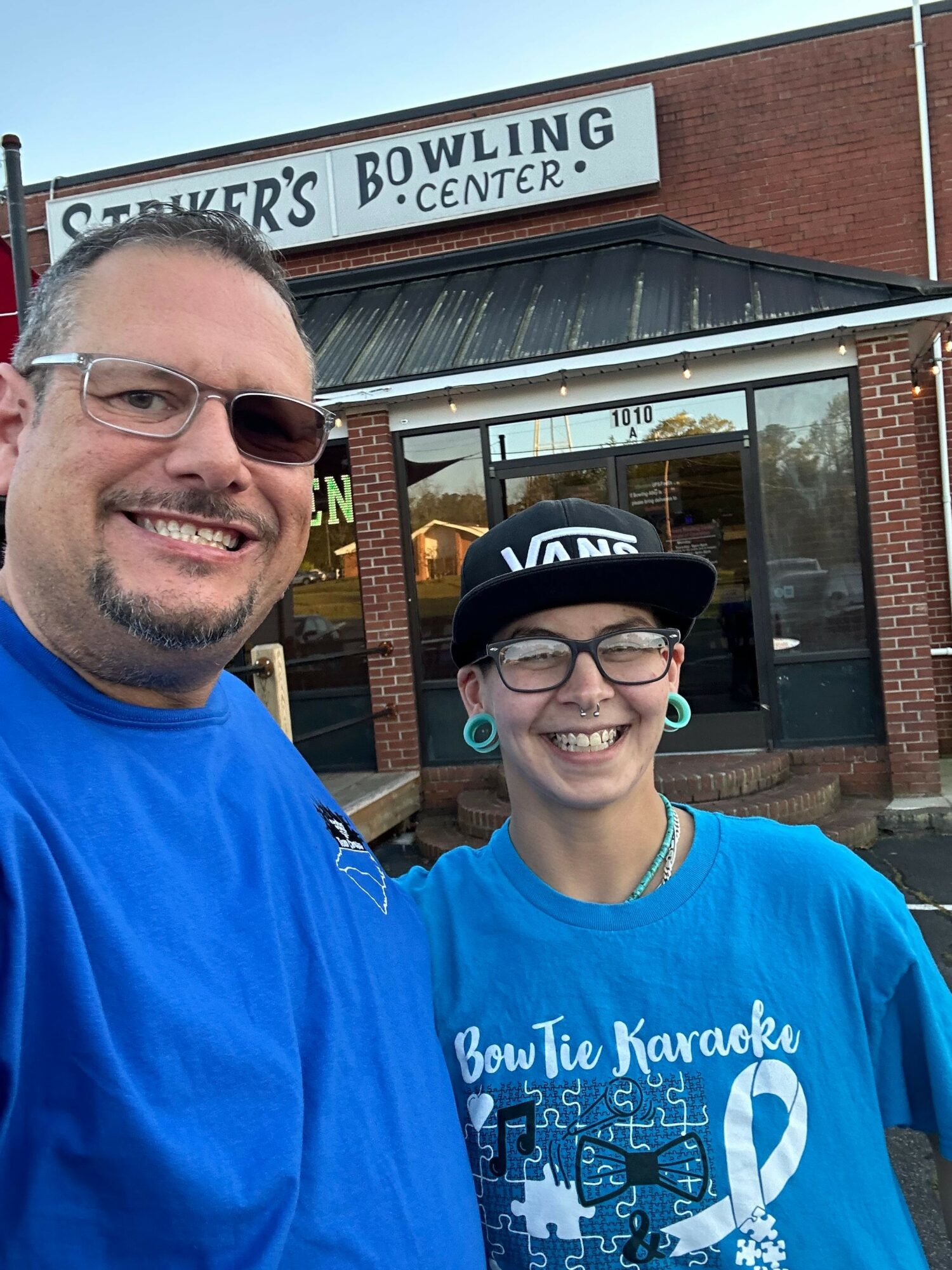 Two smiling people outdoors in front of a bowling alley, one wearing glasses and a blue shirt, the other wearing sunglasses.