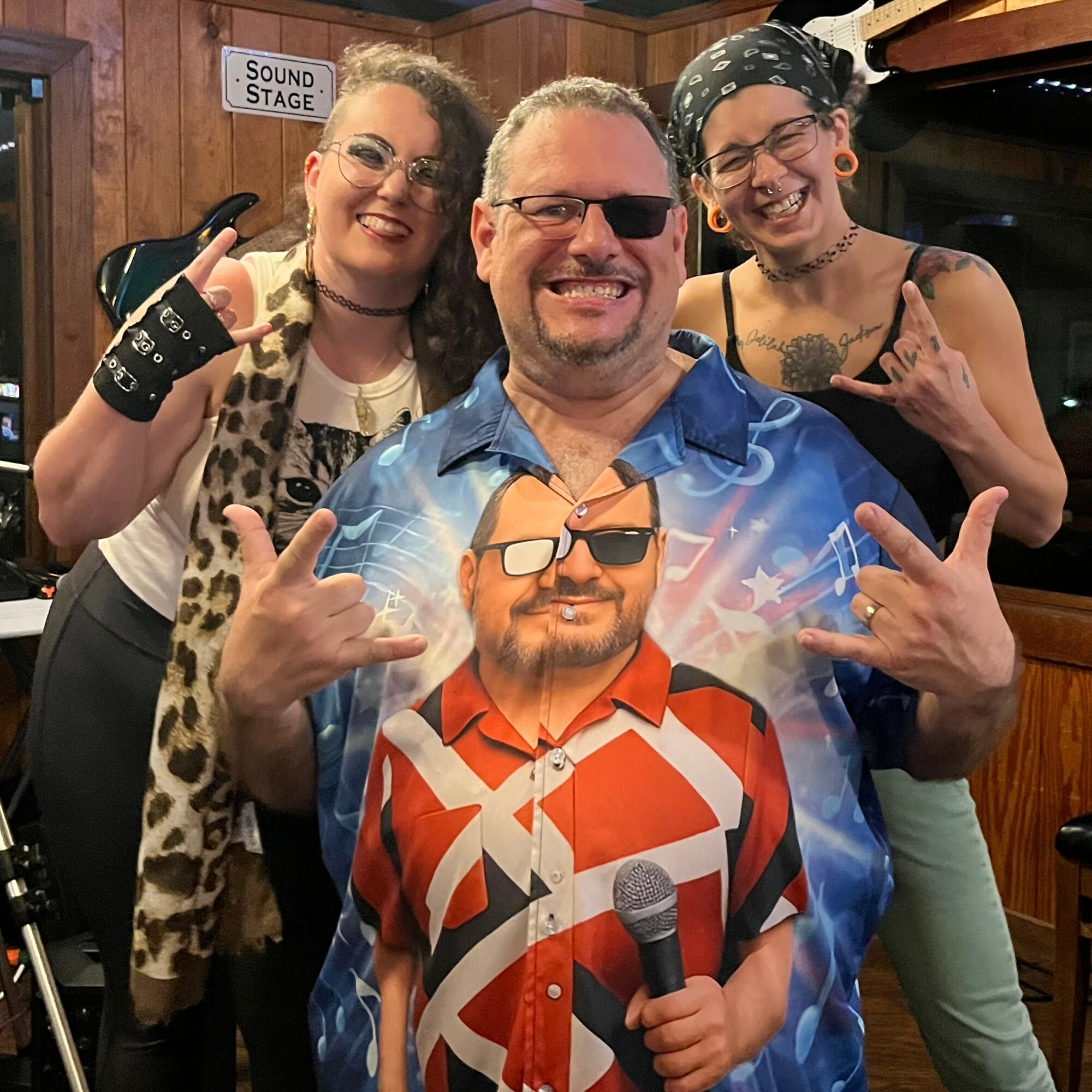 Three people smiling, two women and one man, making rock and roll hand gestures indoors with wooden walls and a sign that says 'Sound Stage'.