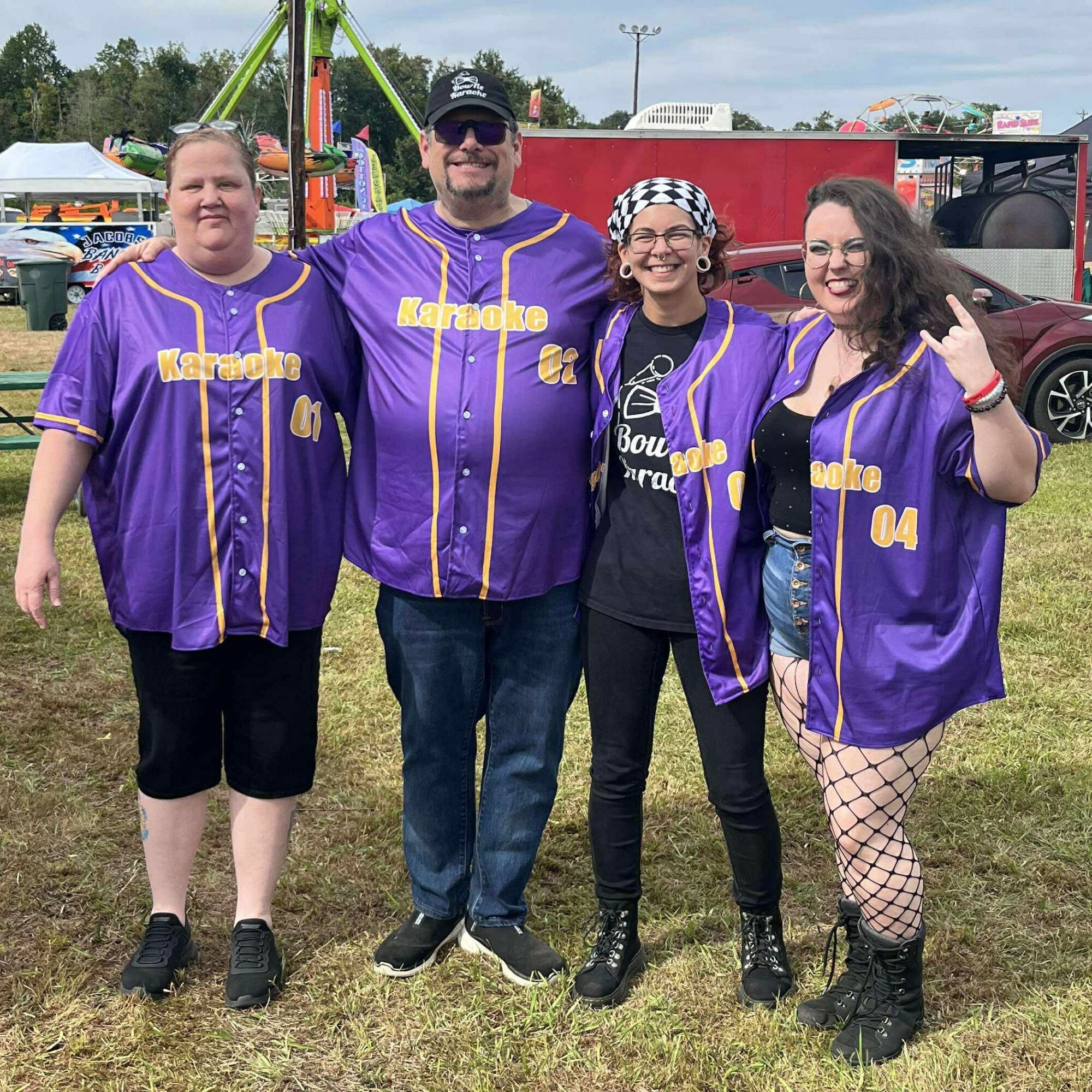 Four people standing outdoors, wearing purple baseball jerseys with yellow accents, smiling, with carnival rides and tents in background.