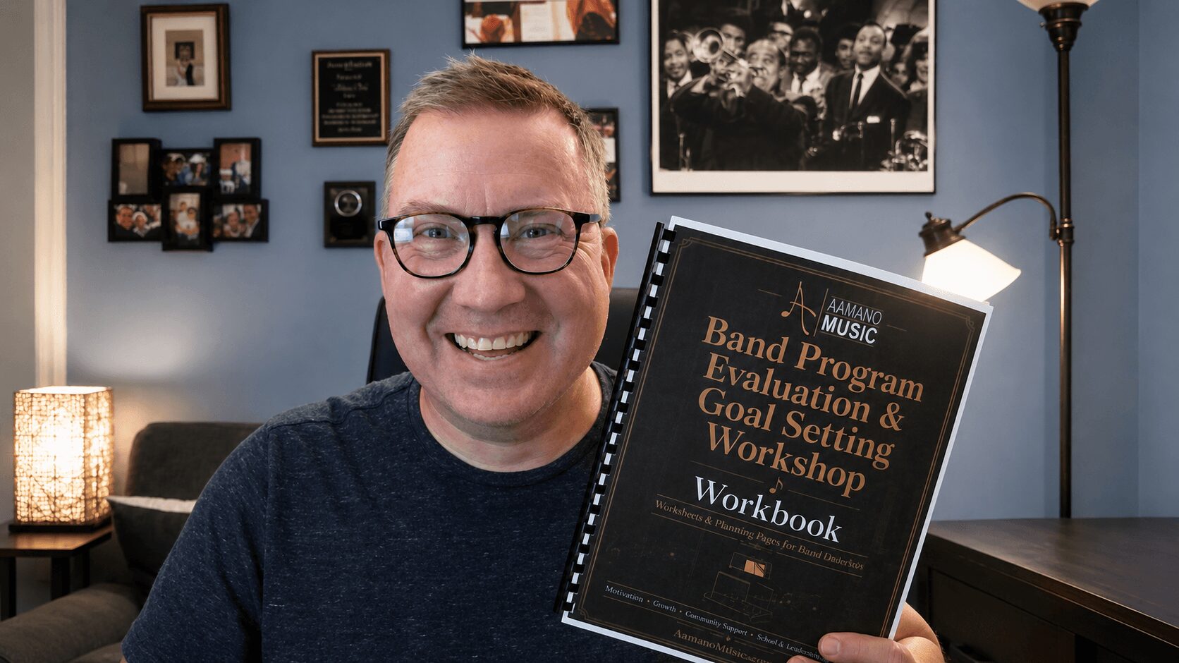 Smiling man with glasses holding a workbook titled 'Band Program Evaluation & Goal Setting Workshop' in a room with framed photos on the wall.