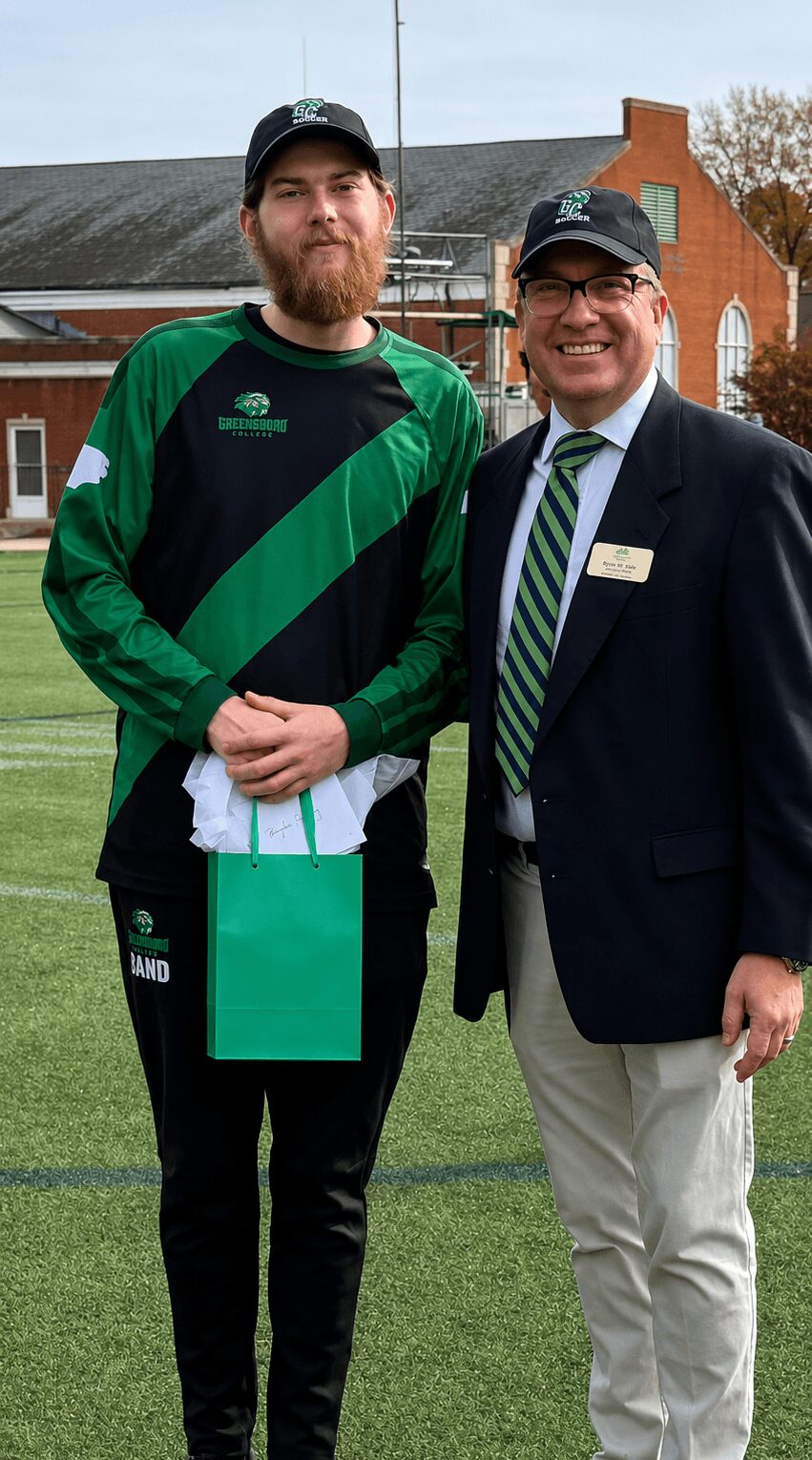 Two men standing outdoors on grass, one in sportswear and the other in a suit, smiling at the camera.