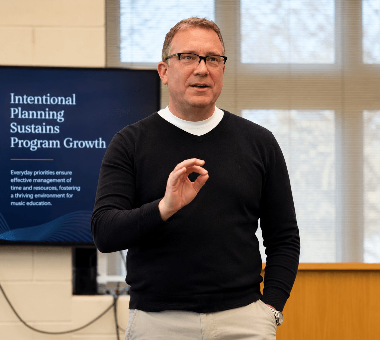 Man in glasses and black sweater speaking in front of a presentation screen, gesturing with his hand, in a room with large windows.