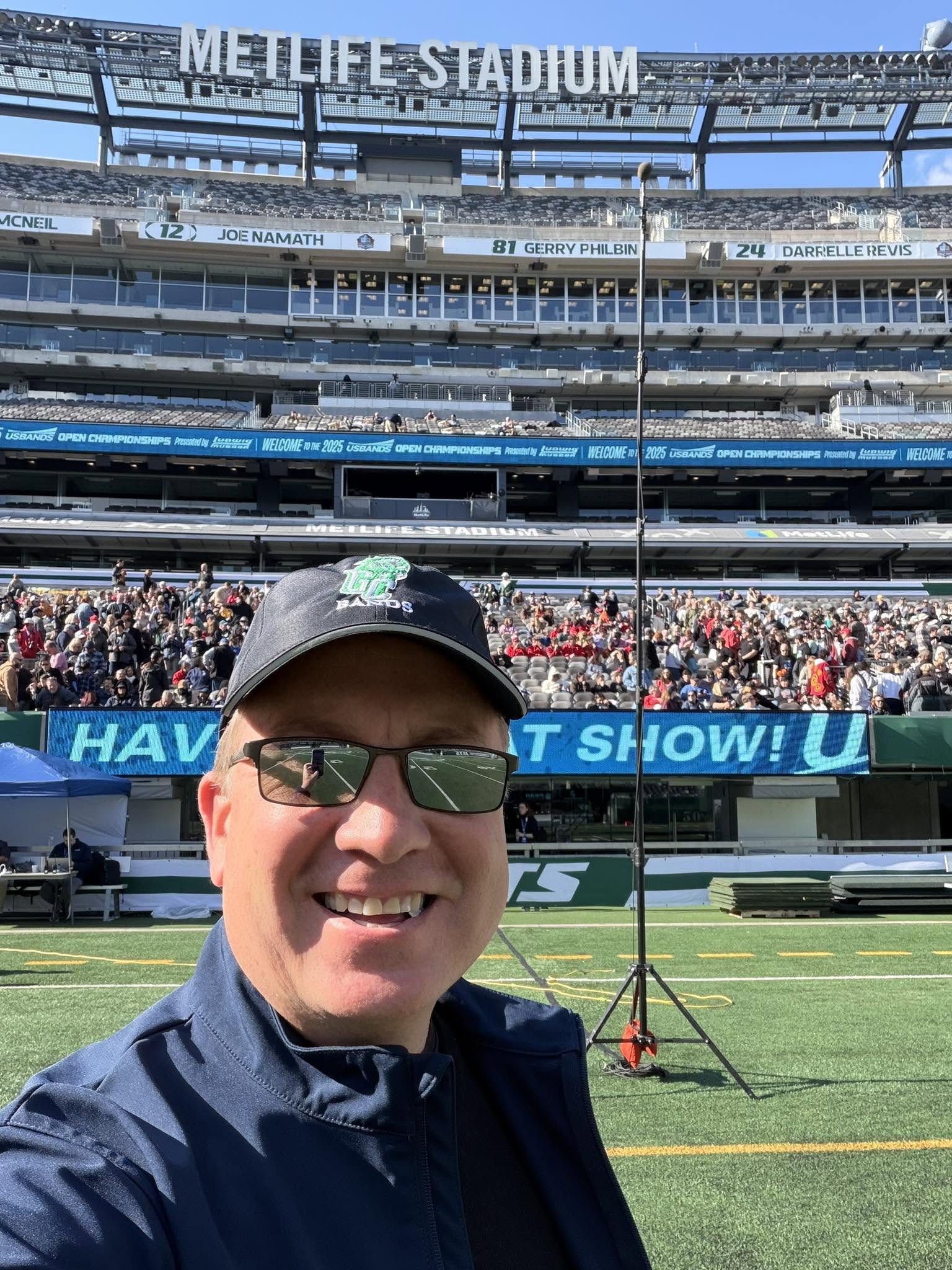Person wearing sunglasses and a cap smiling at a stadium with a crowd and field in background.