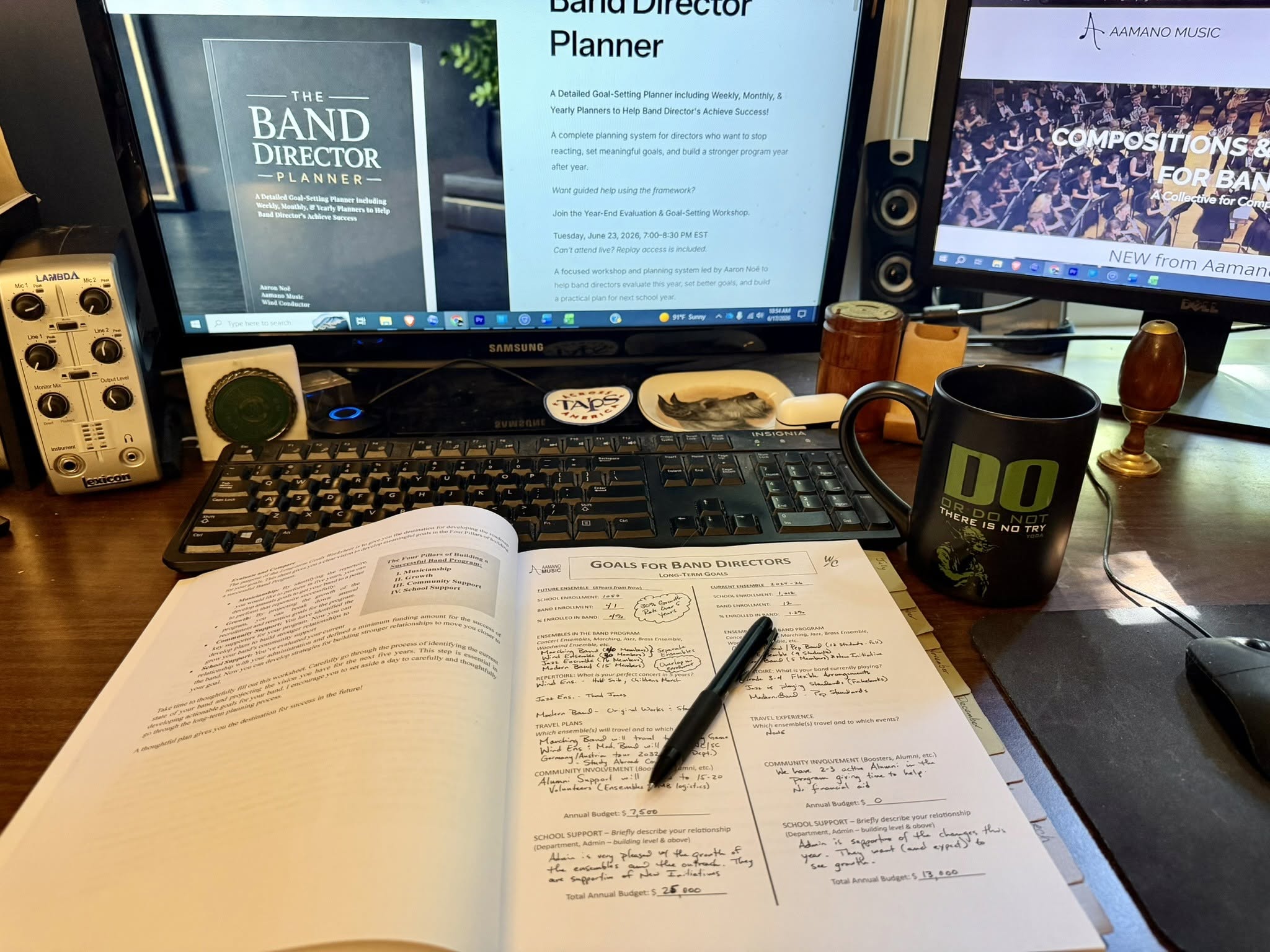 Open book, pen, computer monitors, keyboard, mug, and various items on desk.