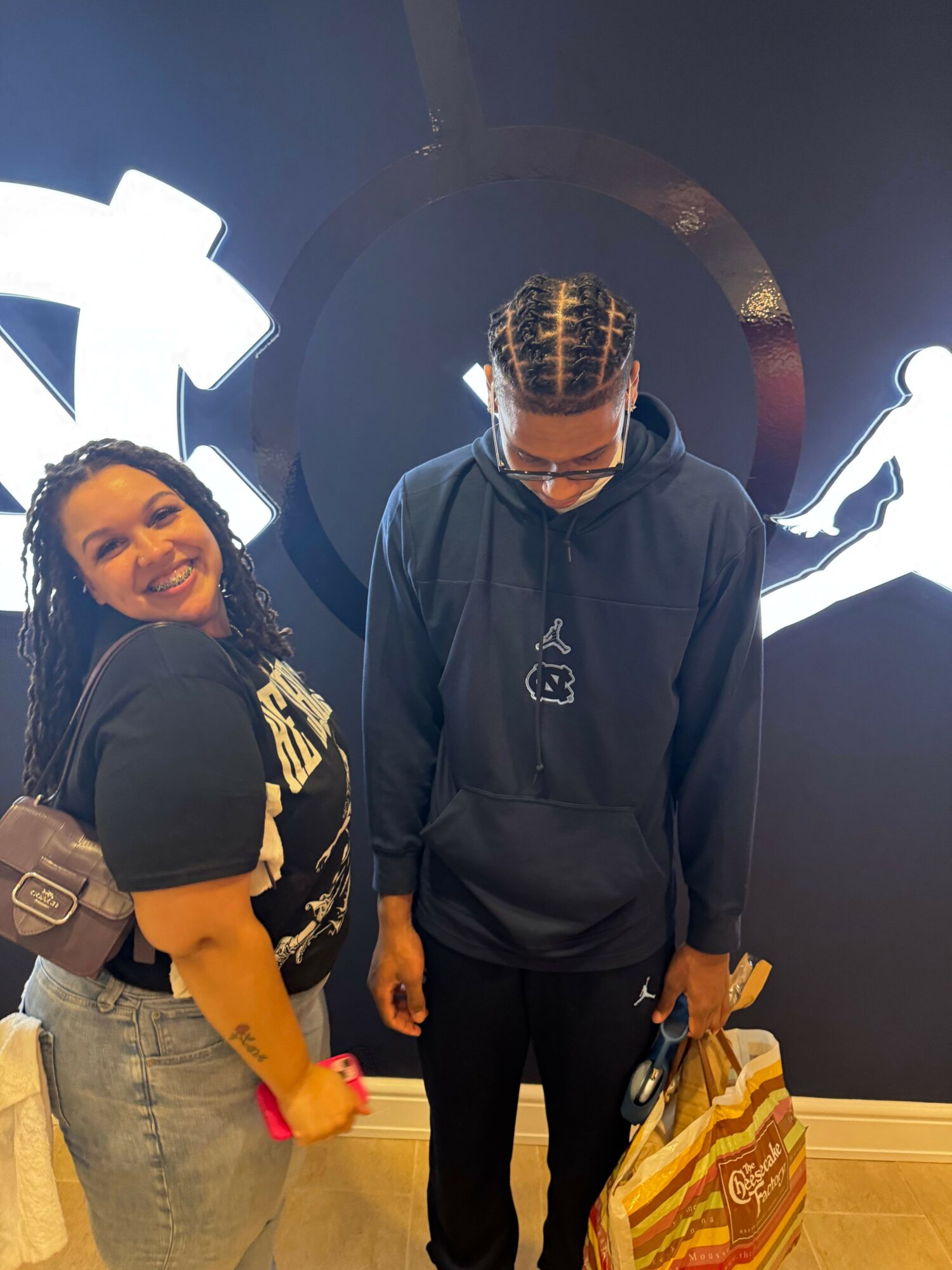 Person with patterned hair and glasses standing in front of a dark background, wearing a black hoodie, with a person in front smiling, holding a red object.
