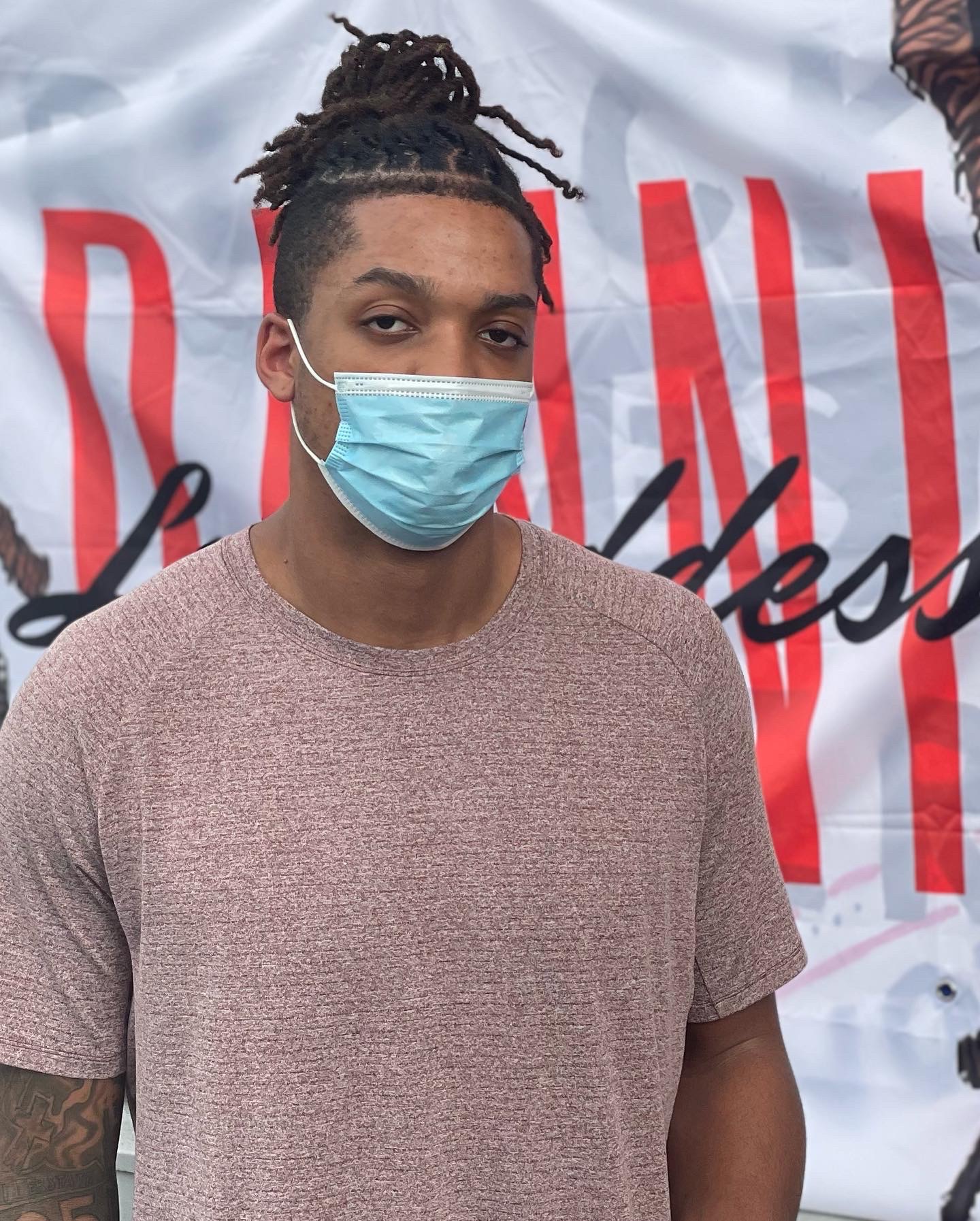 Young person with dreadlocks wearing a face mask and a beige shirt, standing in front of a banner.