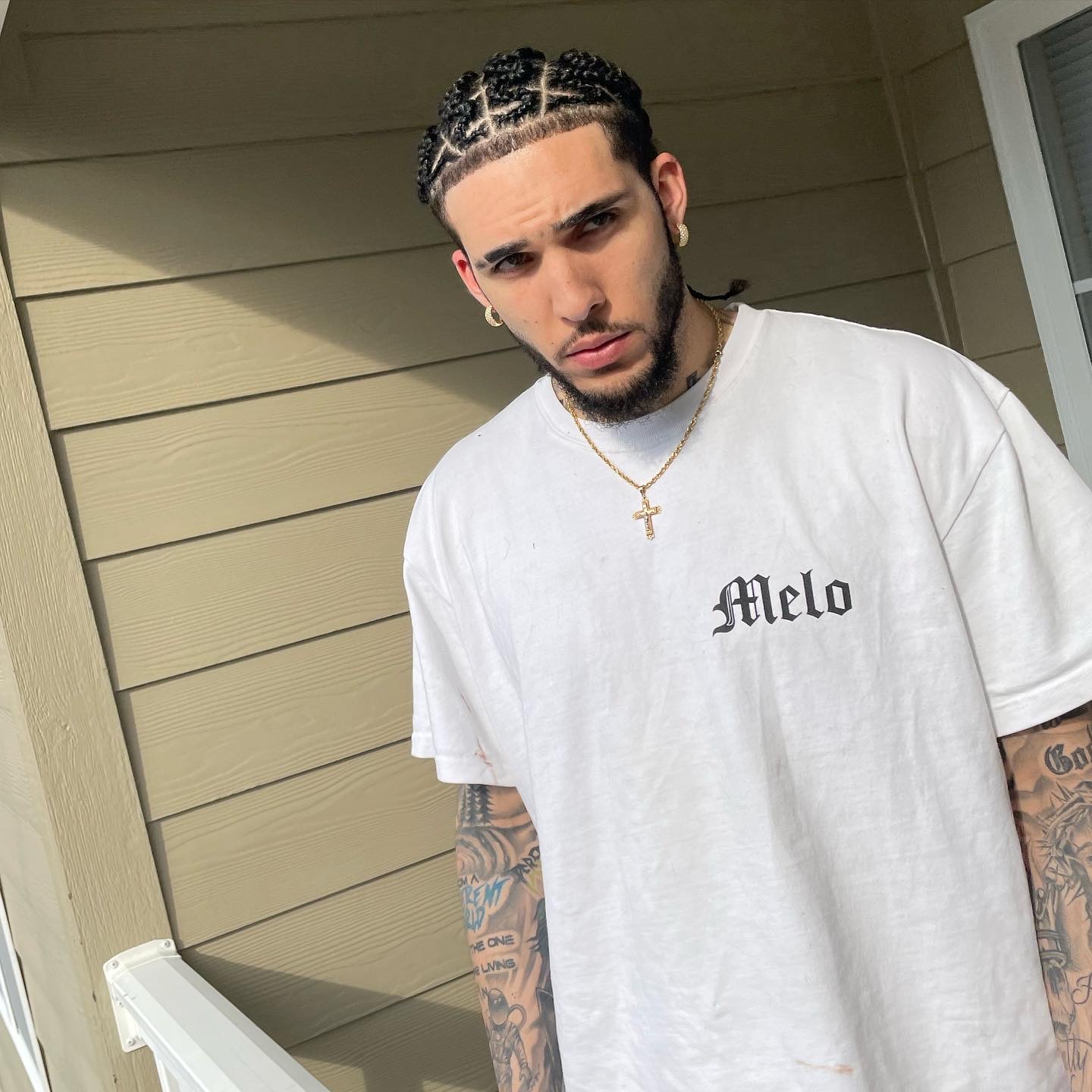 Young man with braided hair and tattoos, wearing a white T-shirt with 'Melo' text, standing outdoors against a beige wall.