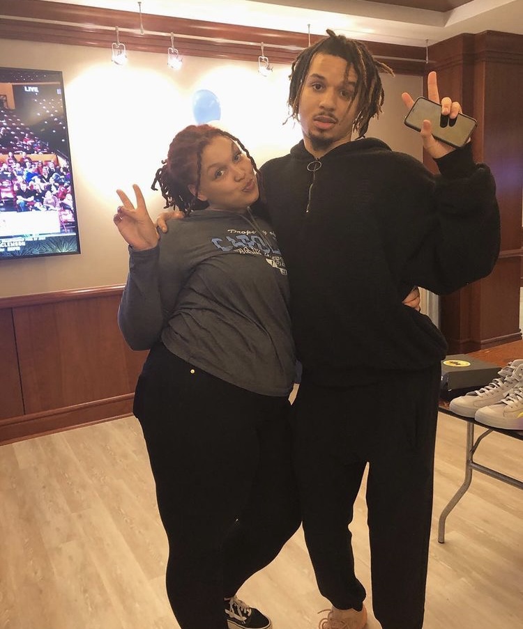 Two people posing indoors, one making a peace sign, the other holding a phone, with a TV screen in the background.