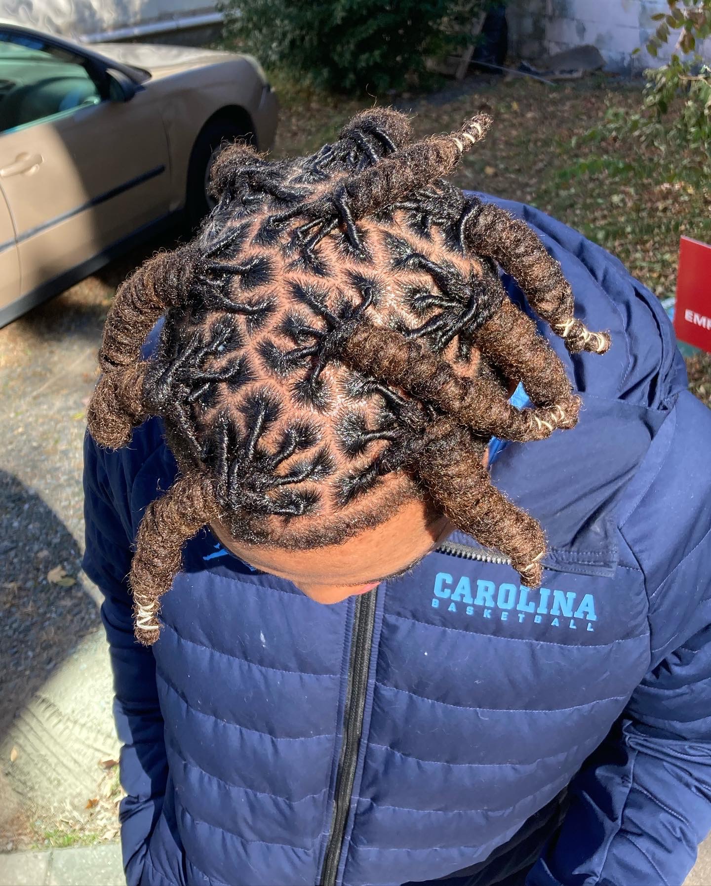 Person with styled dreadlocks outdoors, wearing a blue jacket with 'CAROLINA' text, head bowed down, background includes a car and trees.