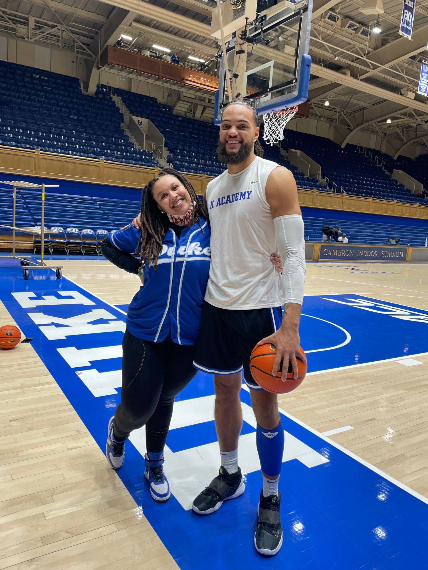 Two women standing on a basketball court, one holding a basketball, with empty blue seats in the background.