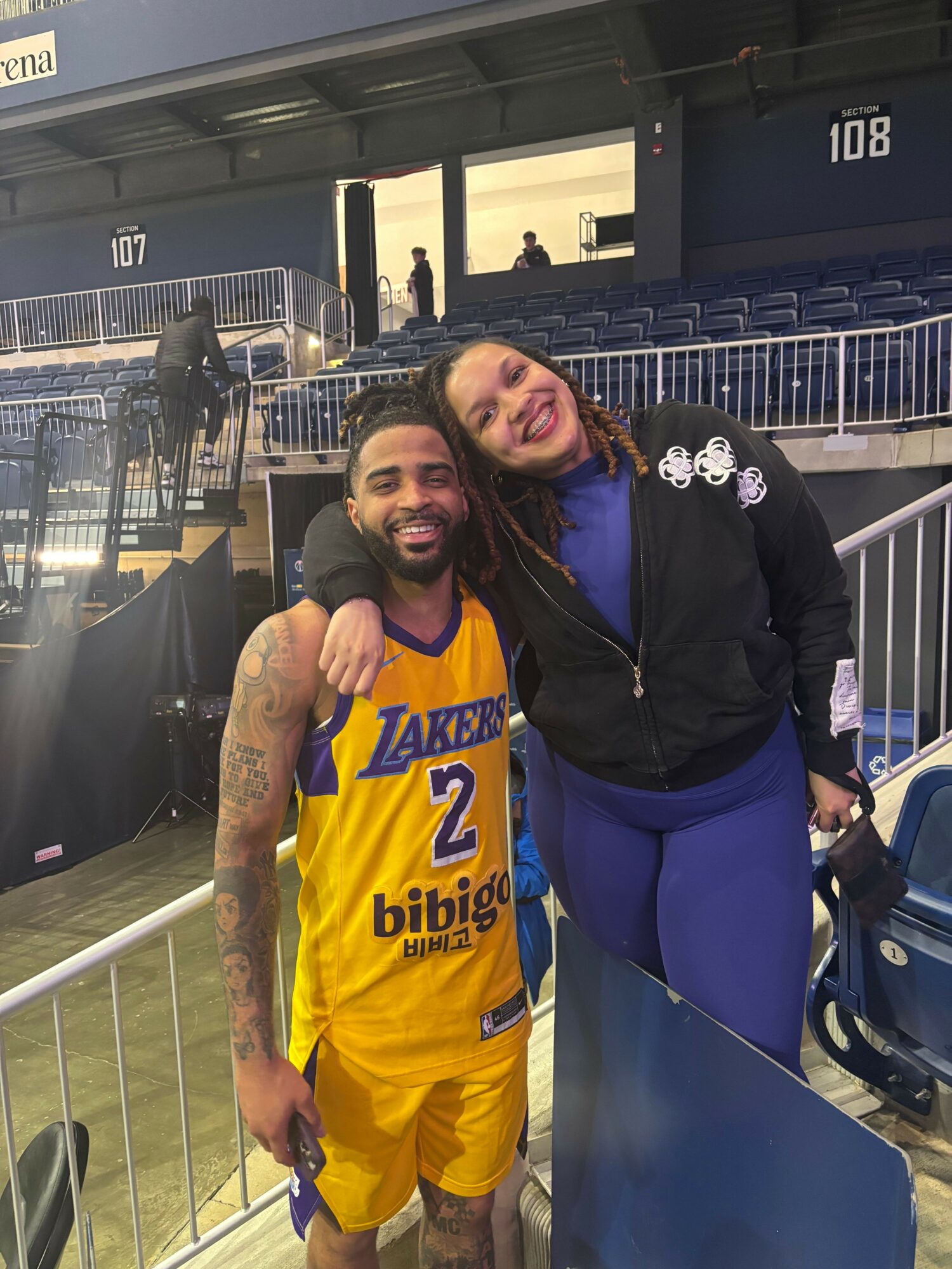 Two women standing at an outdoor event, one in a basketball jersey and the other in a jacket, smiling.