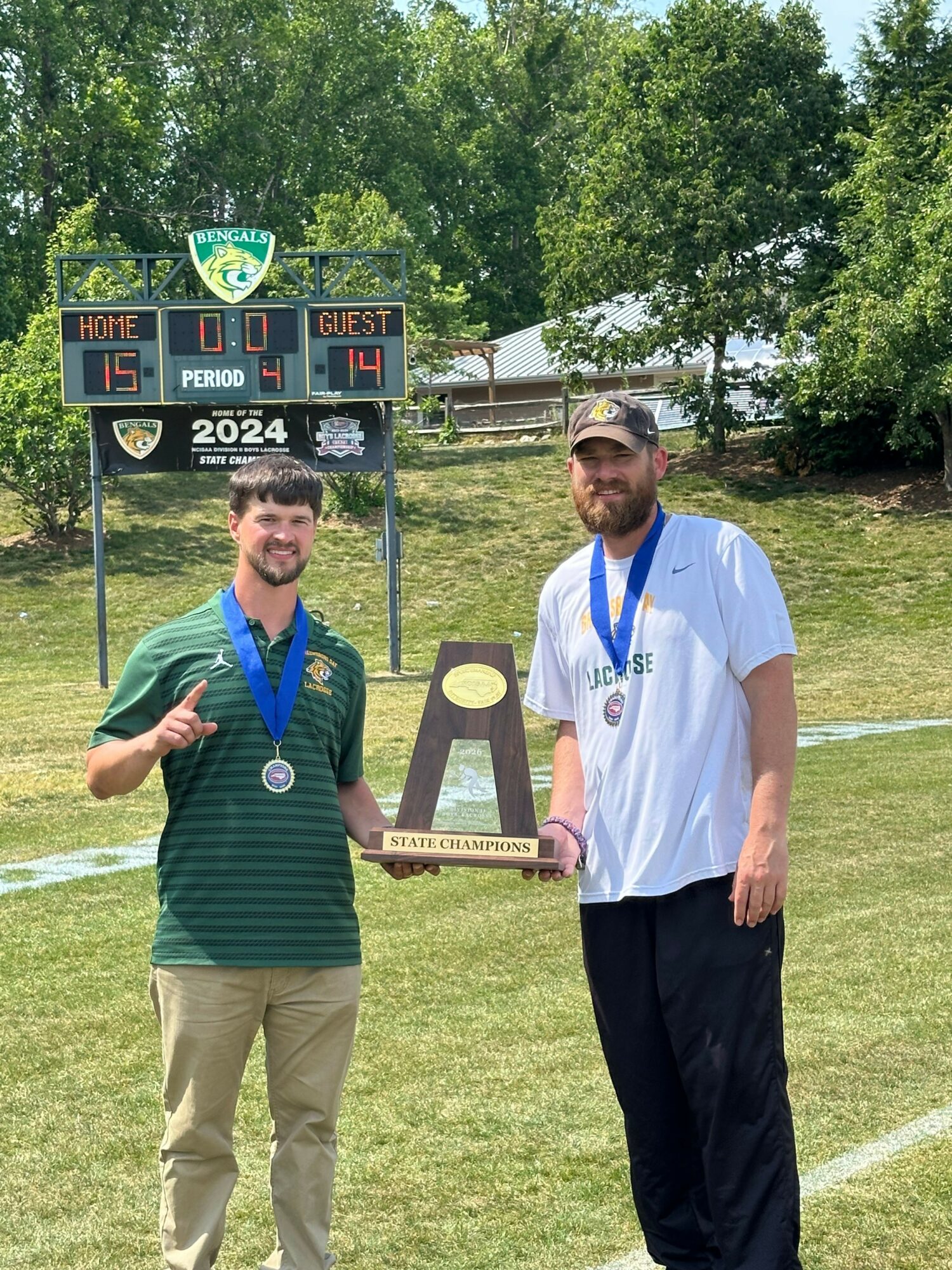 Two men standing outdoors on grass, one holding a trophy, the other giving a thumbs-up, with a scoreboard in the background.