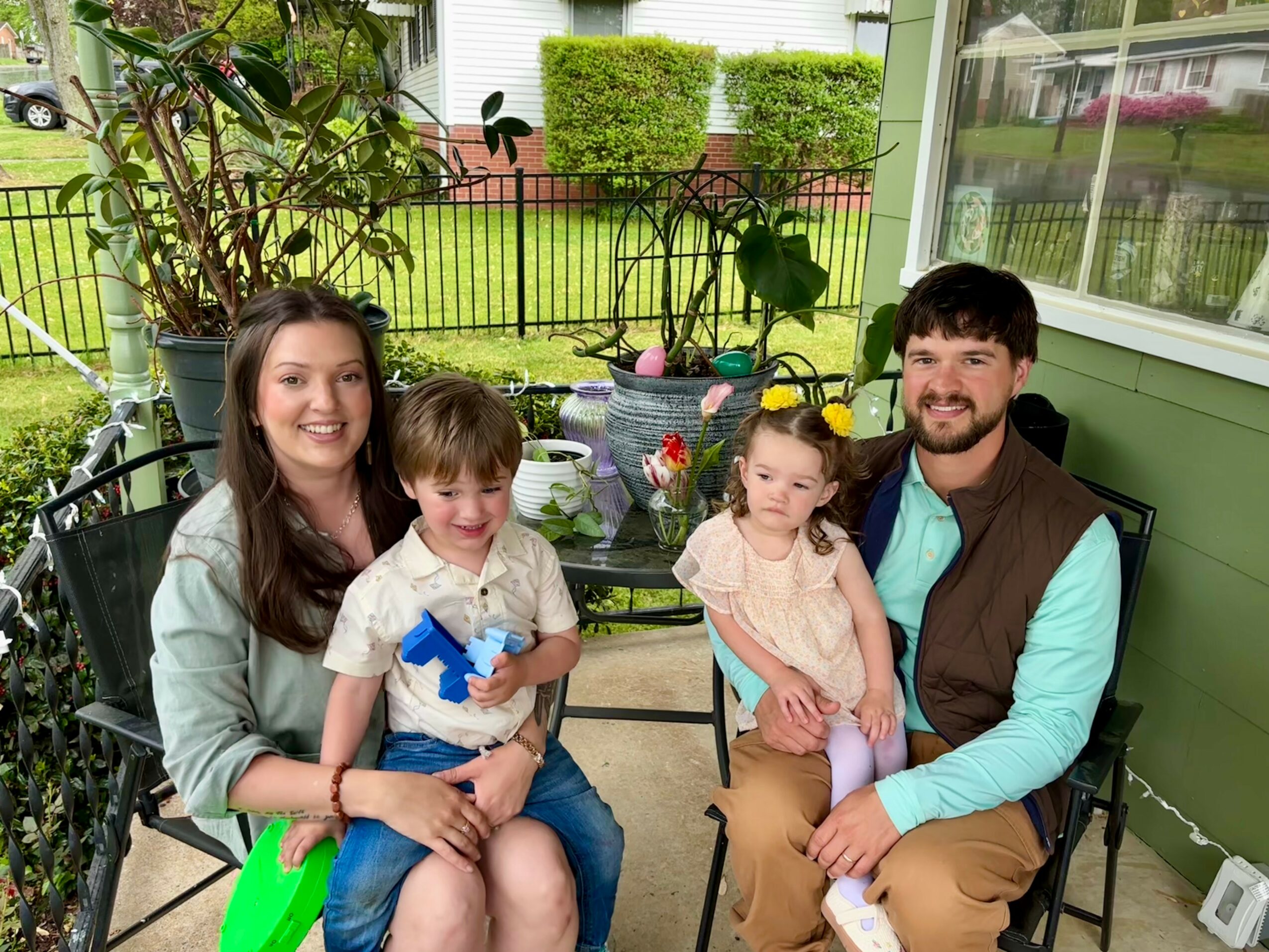 Family of four sitting on a porch with plants and a garden in the background, smiling at the camera.