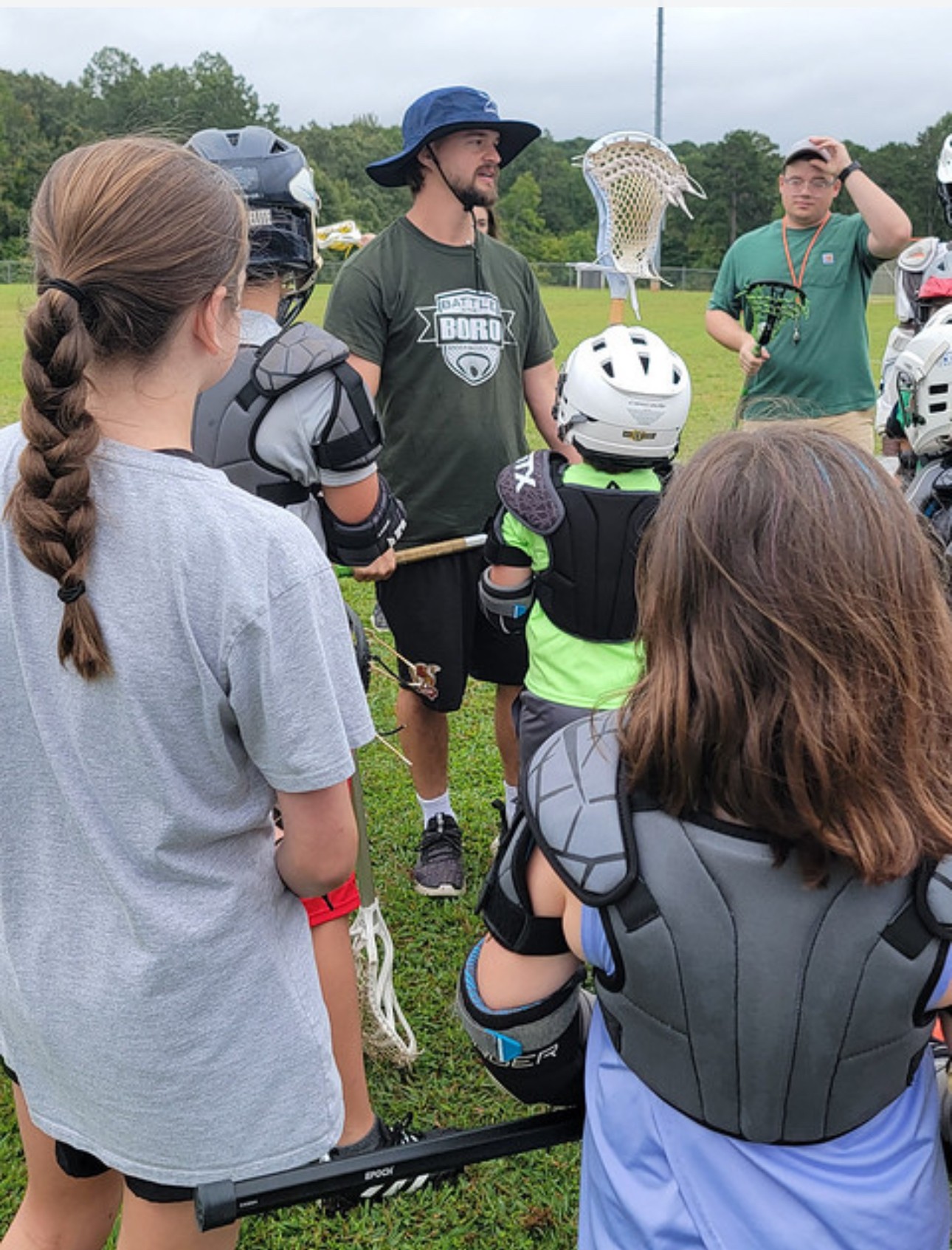 Group of children and a coach outdoors, children wearing lacrosse gear, coach speaking, grassy field, trees, and cars in background.