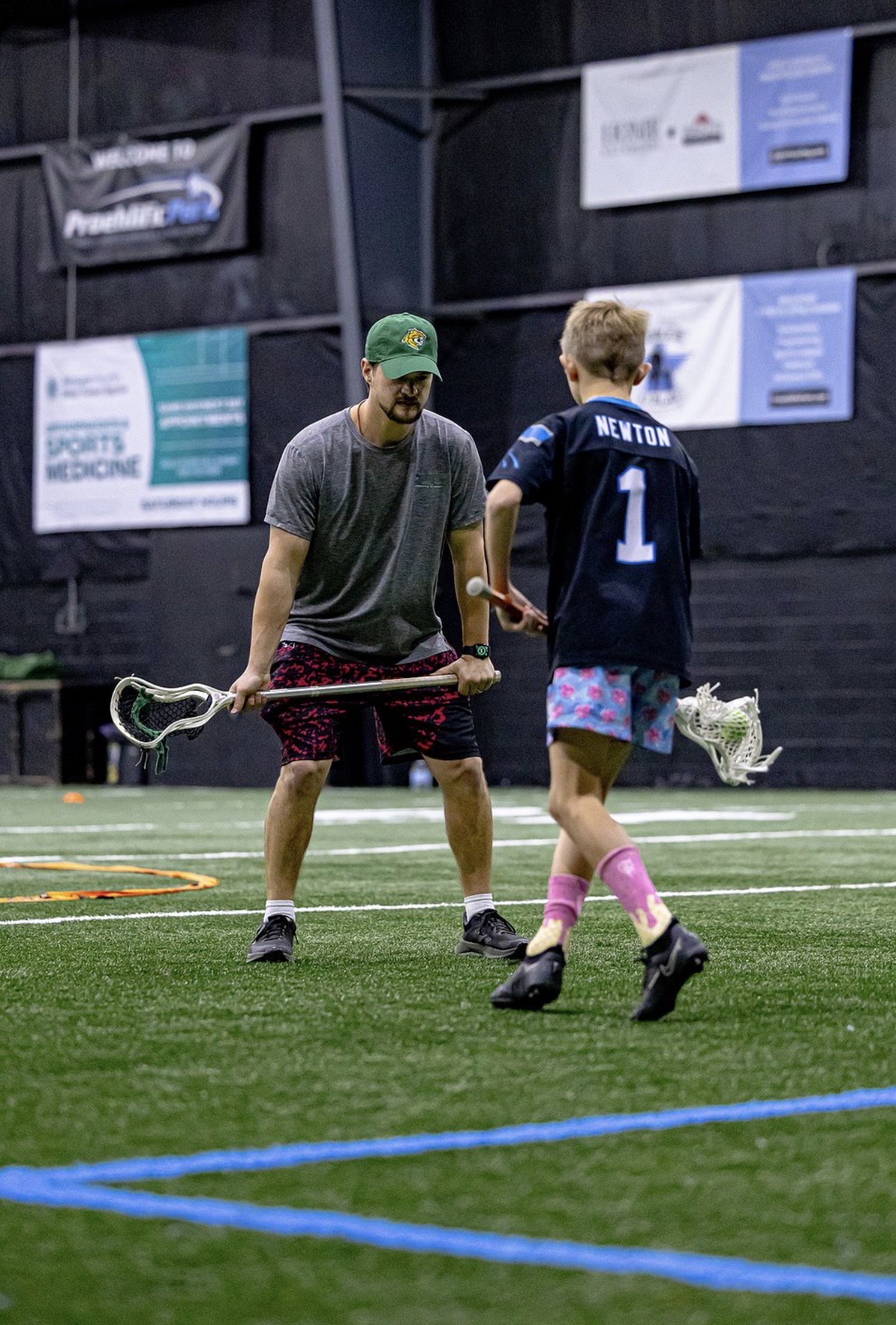 Two people on an indoor sports field, one holding a floorball stick, the other wearing a jersey with the number 1, talking.