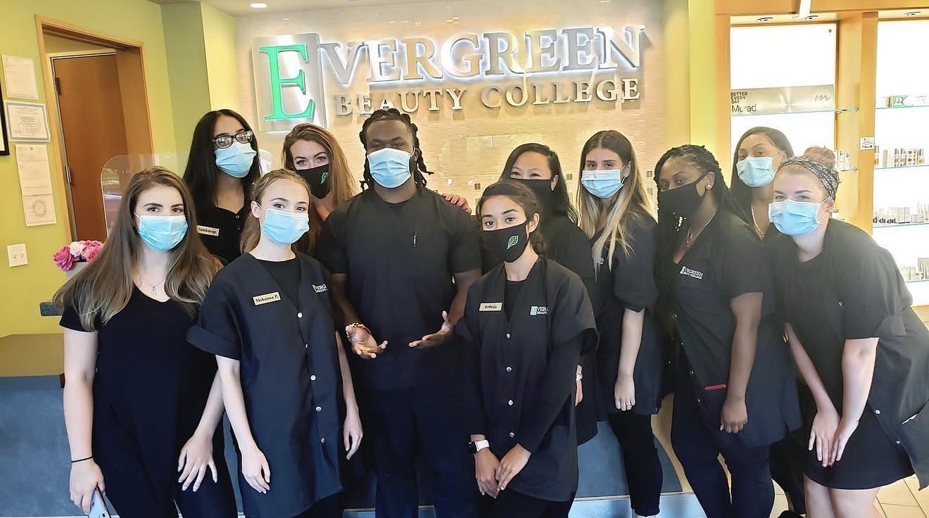 Group of people wearing masks standing inside a building with a sign reading 'Evergreen Beauty College' behind them.
