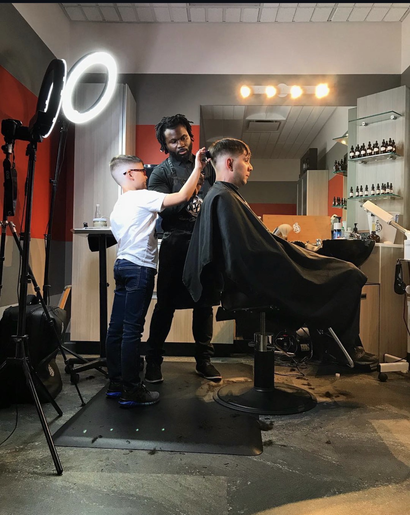 Barber cutting a person's hair in a salon with two children observing, illuminated by ring light, shelves with products in background.