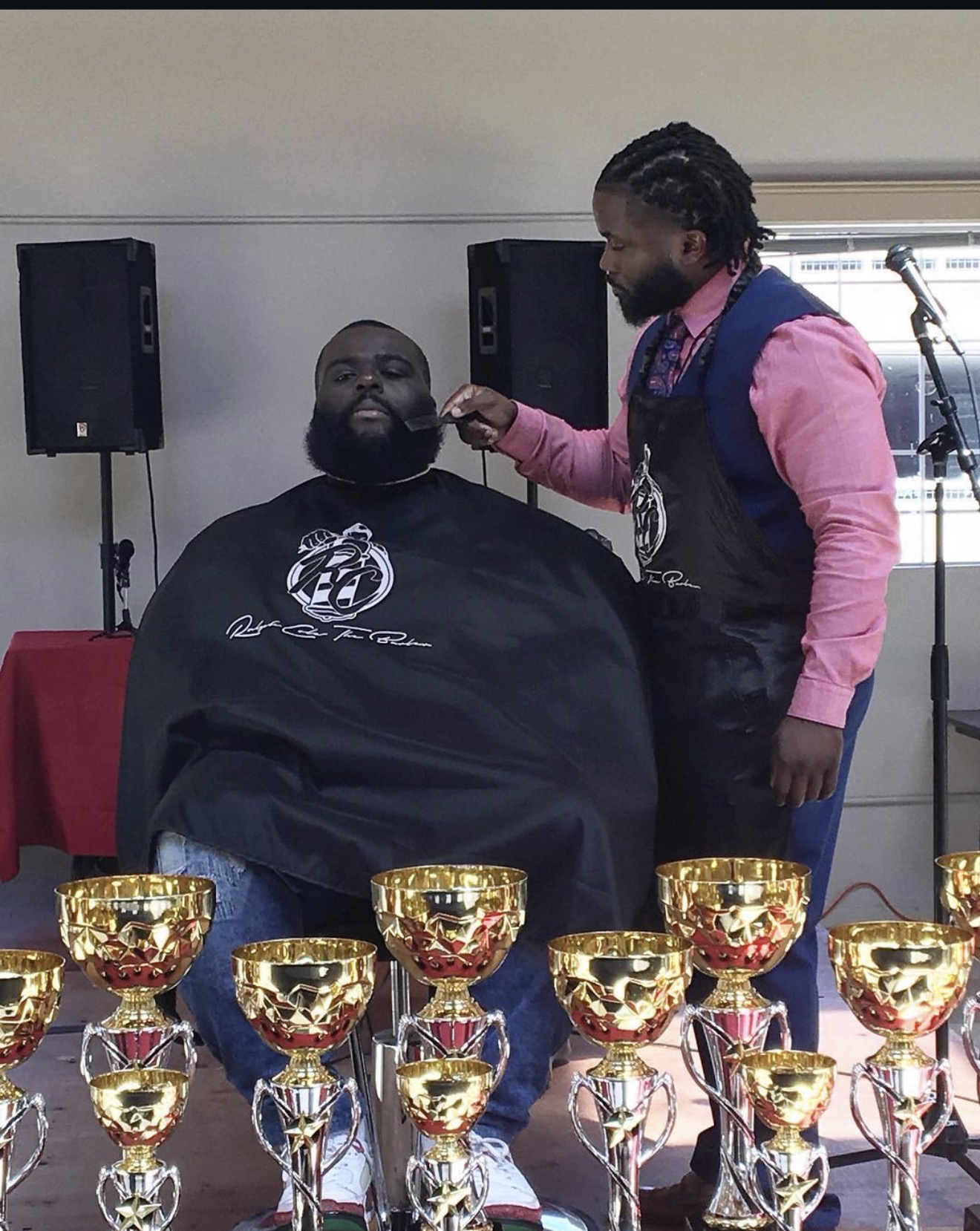 Man receiving a beard grooming or styling service from a stylist in a room with speakers and trophies in the foreground.