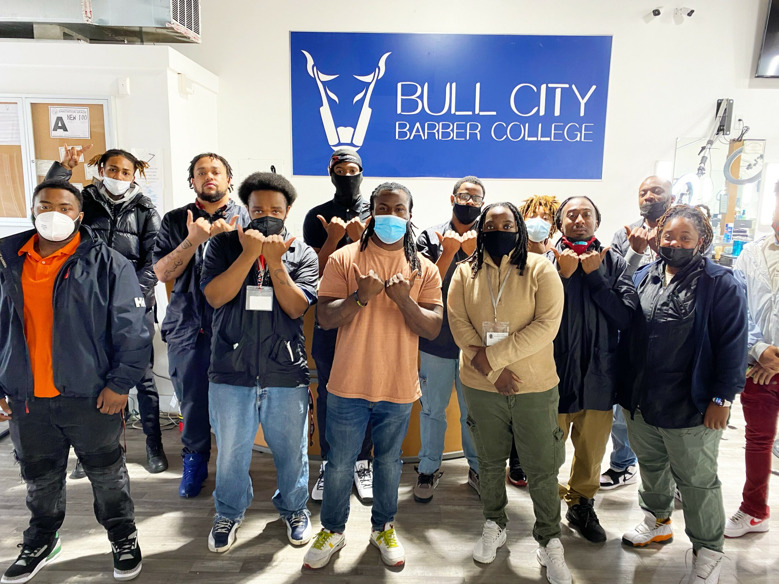 Group of people standing indoors in front of a blue sign that reads 'BULL CITY BARBER COLLEGE', some wearing masks.