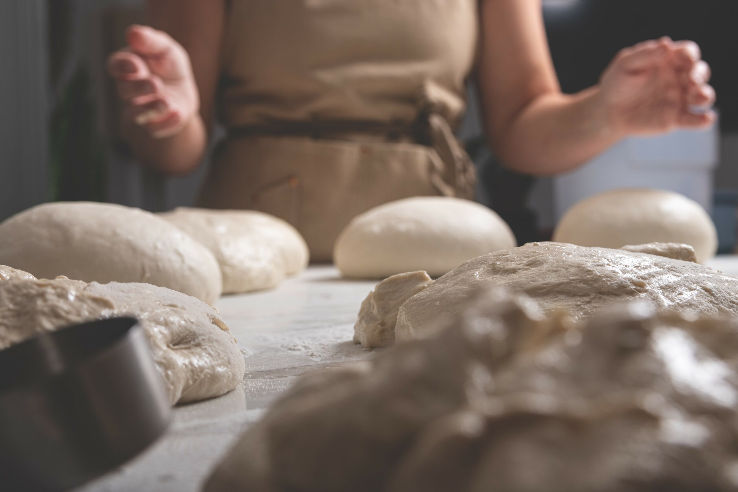 Person in apron working with dough on a table, several dough balls and a rolling pin visible.