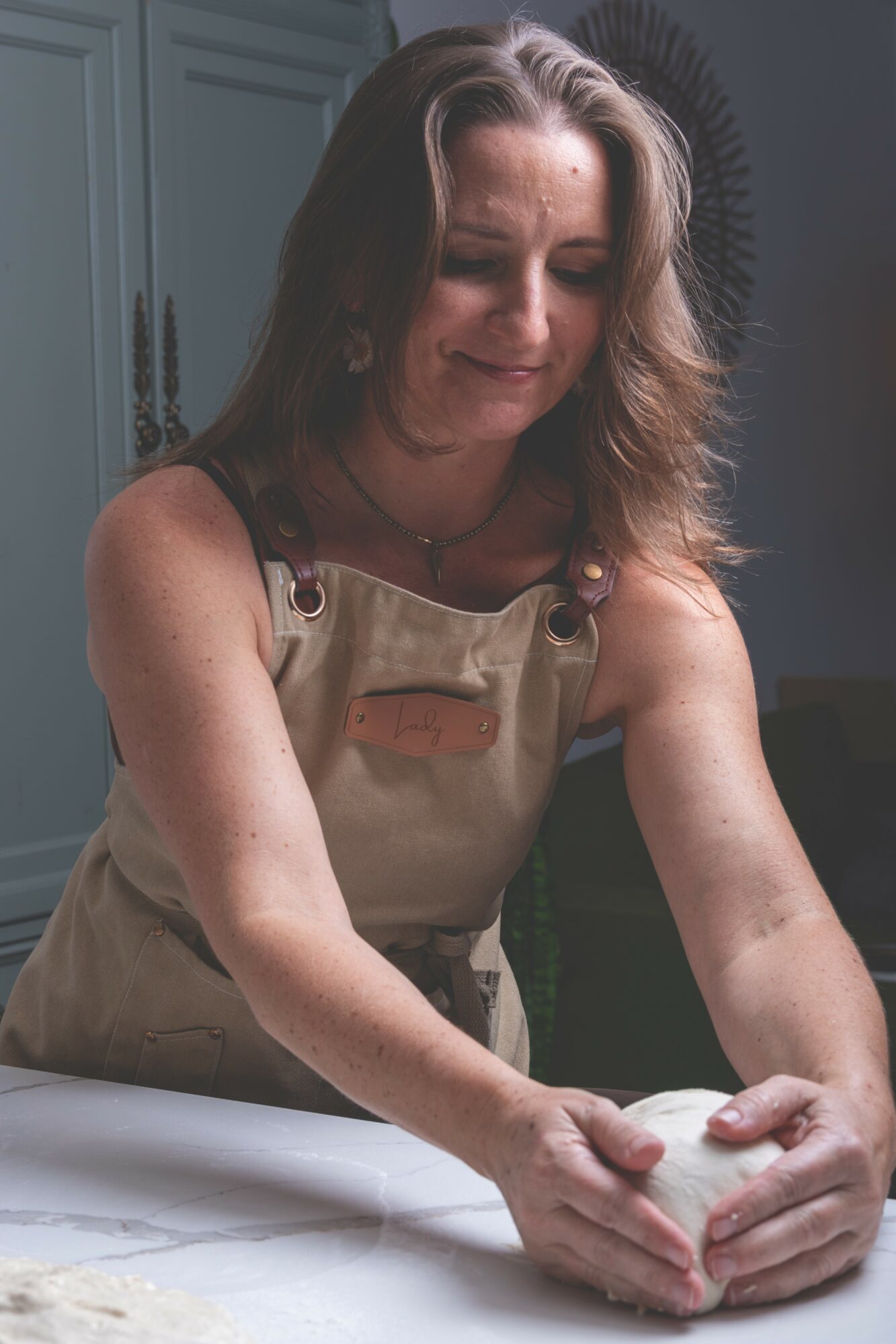 Woman kneading dough on a table, wearing an apron, in a kitchen setting.