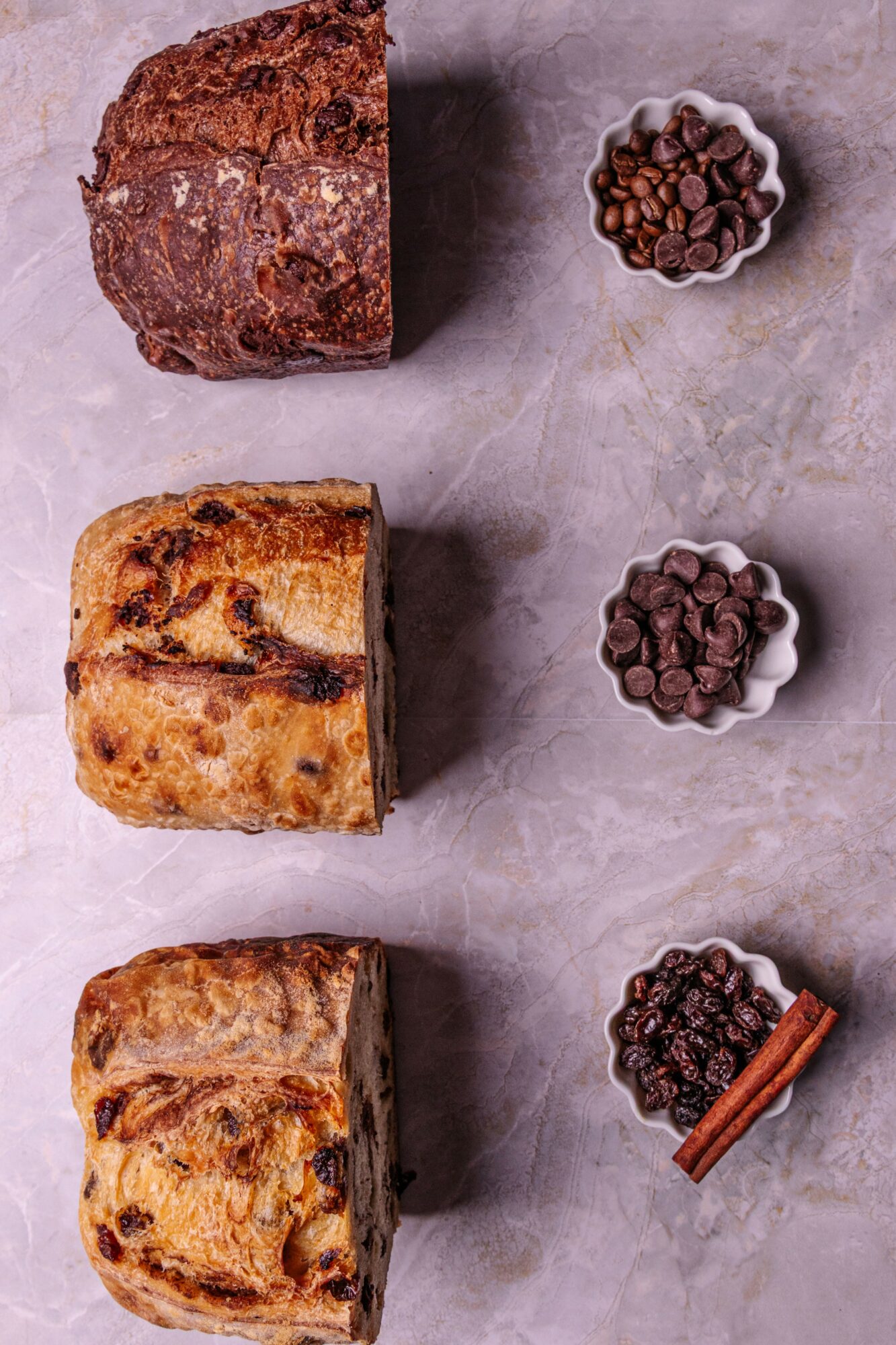 Three slices of cake with chocolate chips and cinnamon sticks, each paired with a bowl of chocolate chips, on a gray surface.
