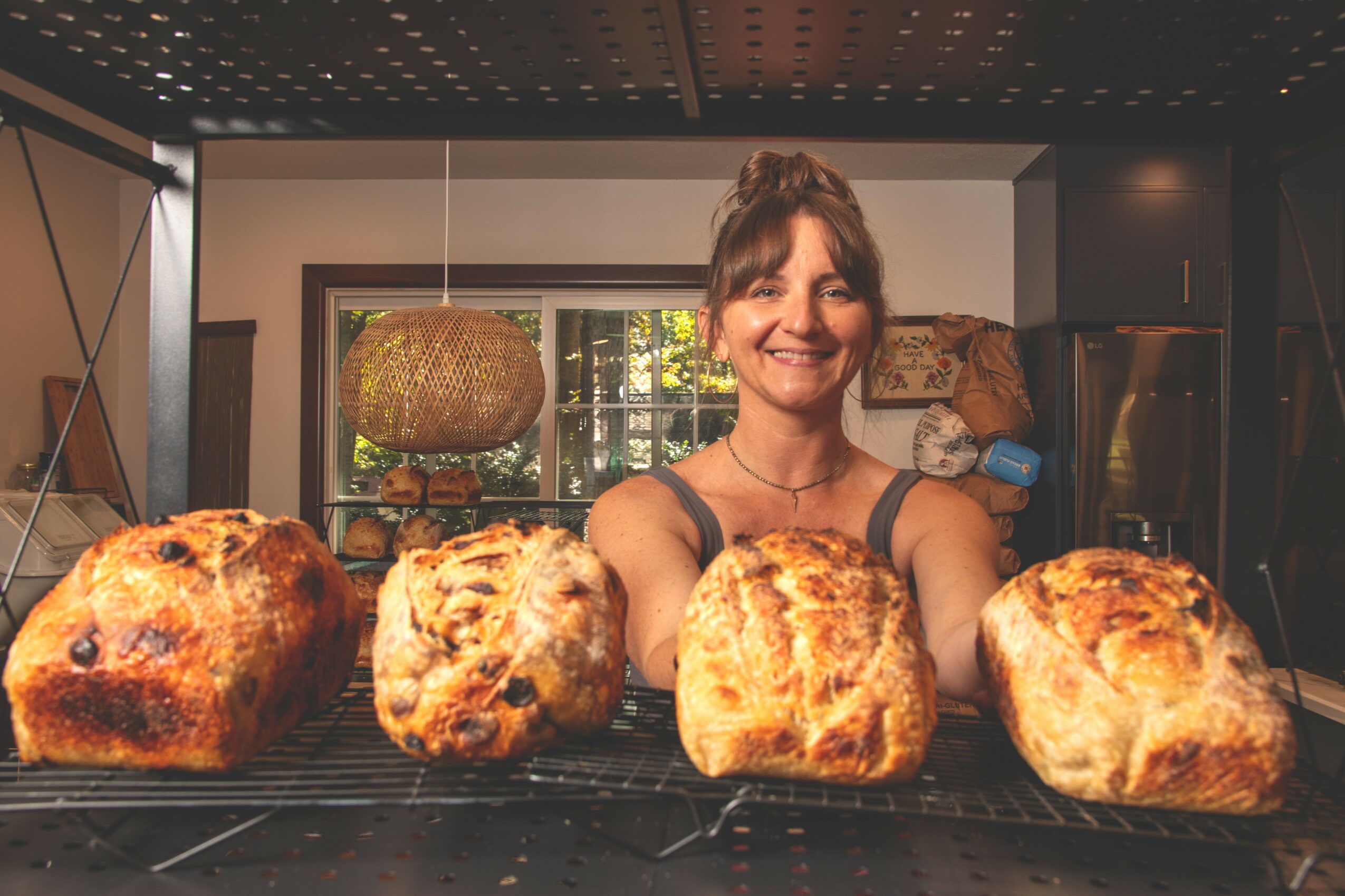 Smiling woman behind four freshly baked loaves of bread on a cooling rack in a kitchen.