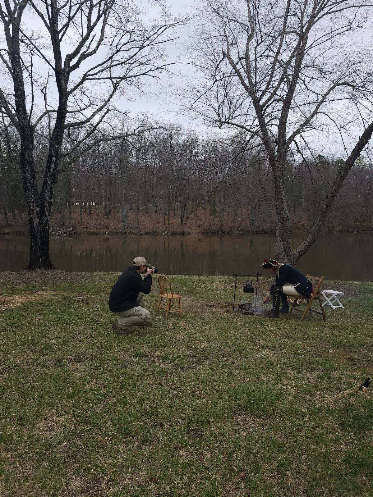 Two people are fishing near a lake with leafless trees in the background, one is kneeling and the other is sitting, with fishing gear around them.