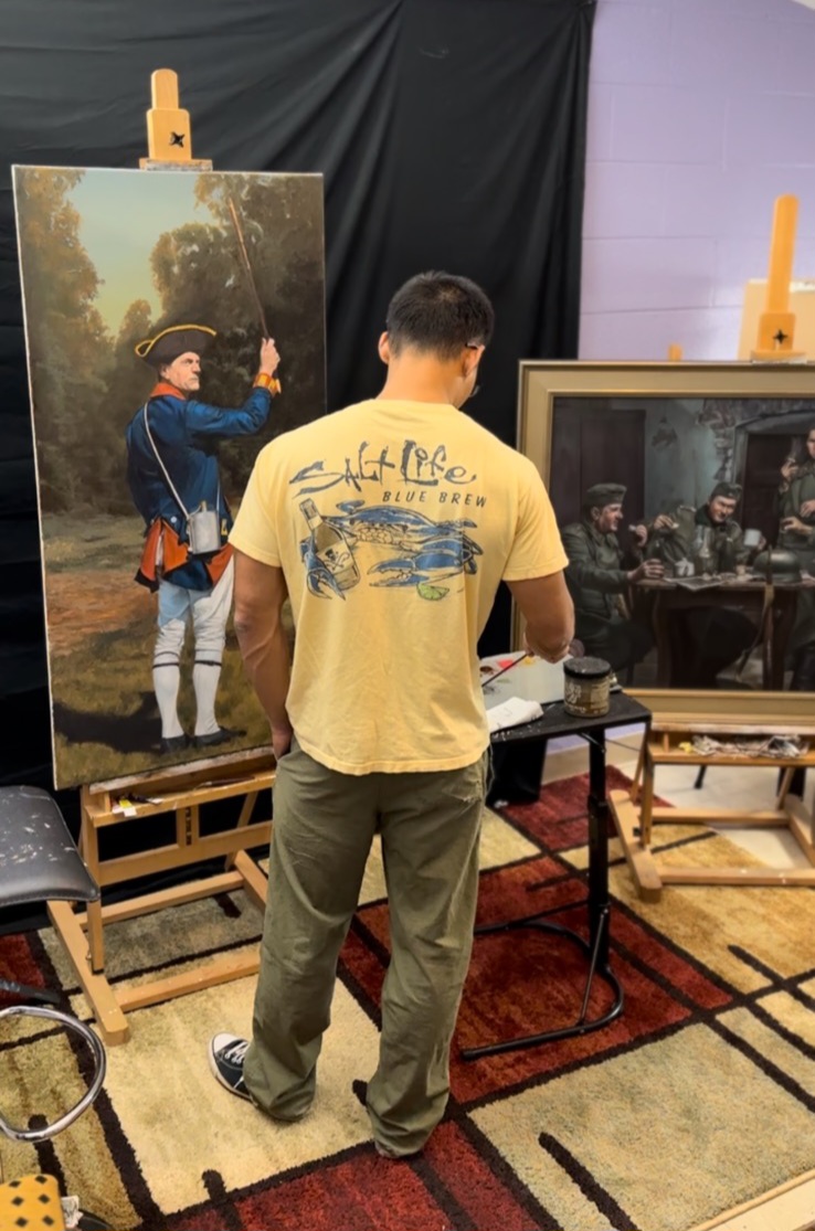 Man with short dark hair wearing a yellow T-shirt and gray pants stands in front of an easel with a painting of a person in a blue jacket outdoors. The scene is indoors with black and purple walls, a small table, and a framed photograph. The man is facing away from the camera.