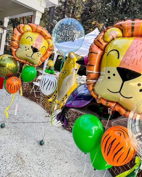 Colorful lion-themed balloons, green and orange balloons, and a blue balloon with text, arranged outdoors on a sidewalk.