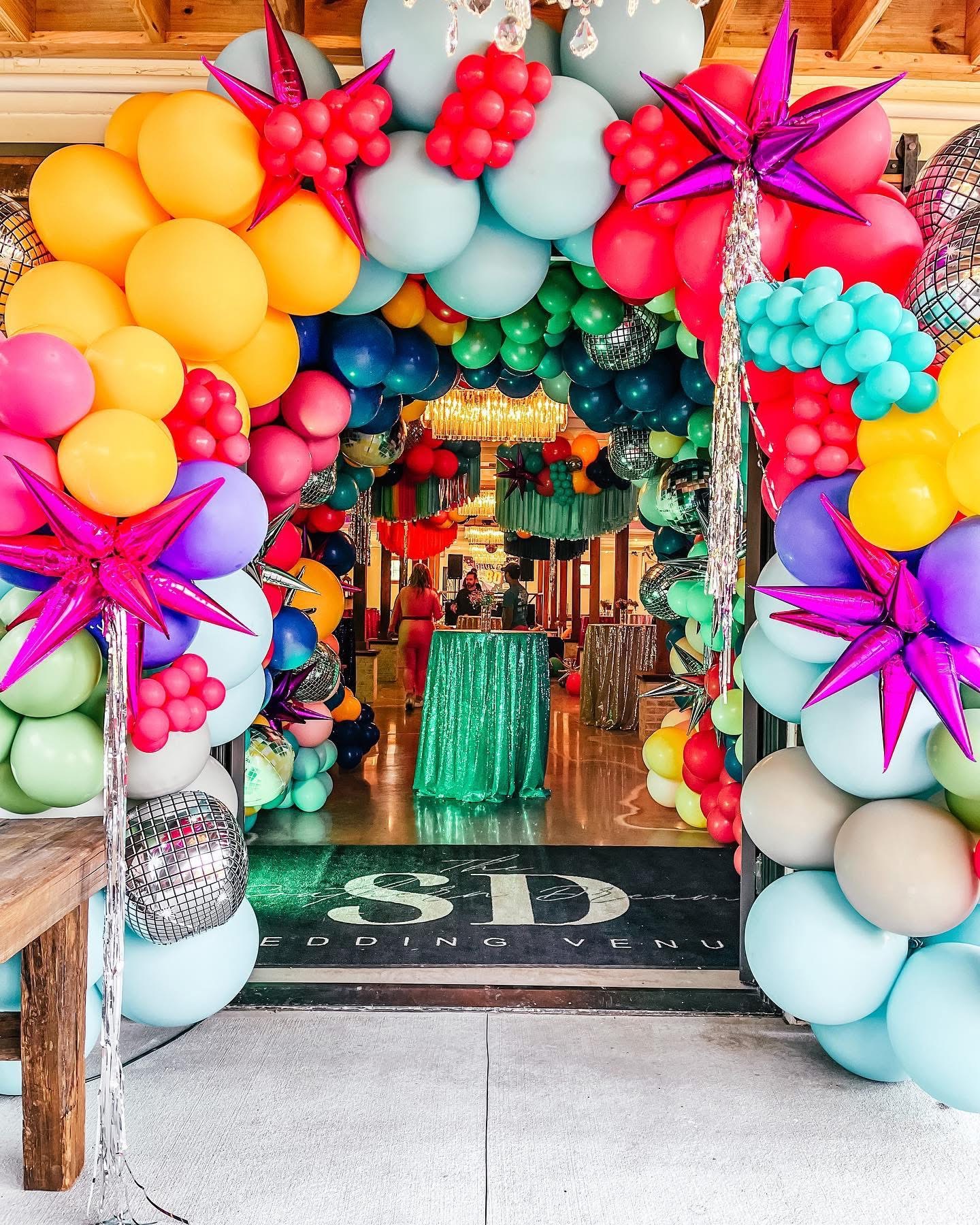 Colorful balloon arch with stars and flowers decorates entrance to event space, with a green table and people inside.