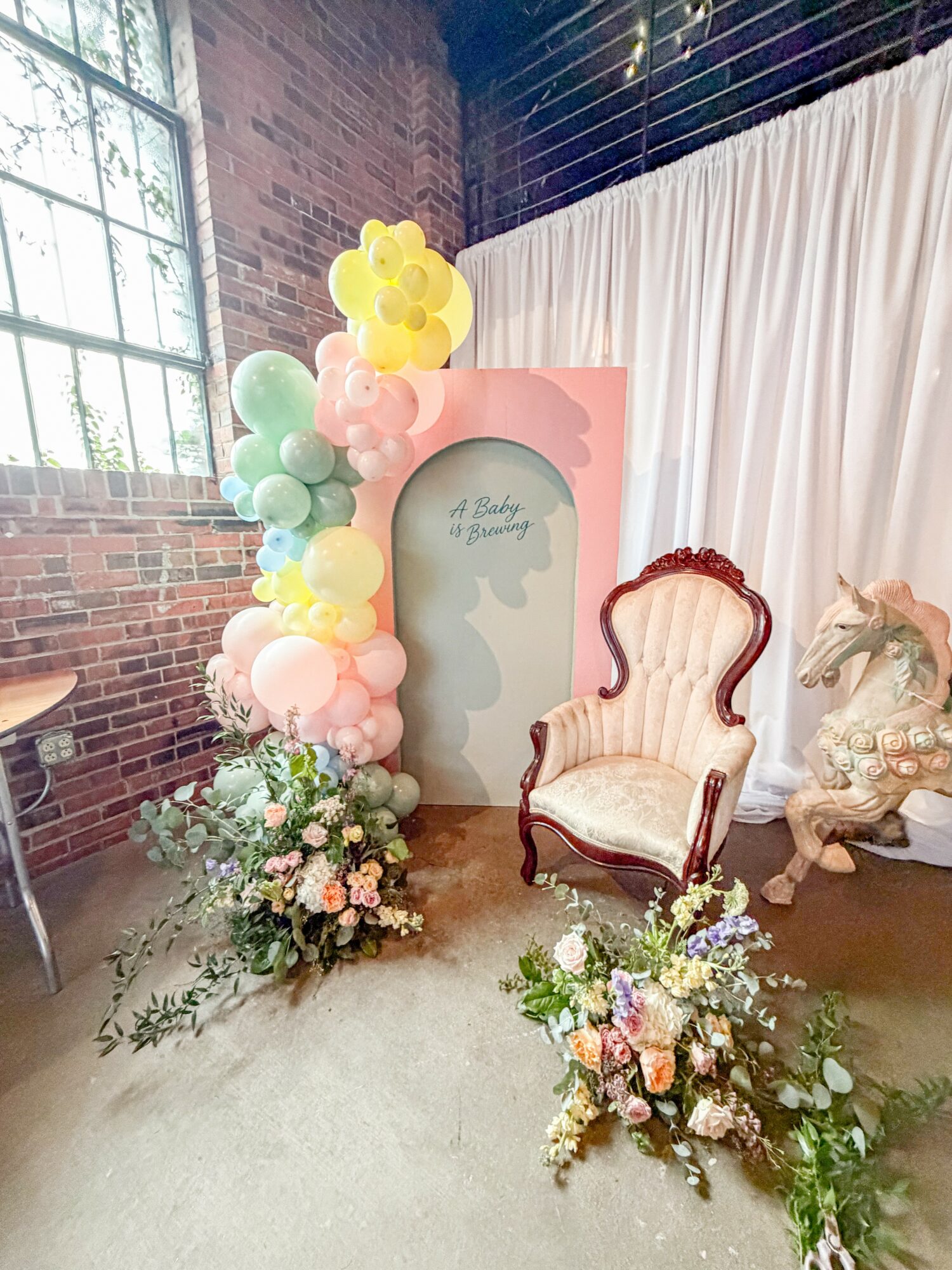 Decorative setup with balloons, floral arrangements, a vintage armchair, and a backdrop with a window and brick wall.