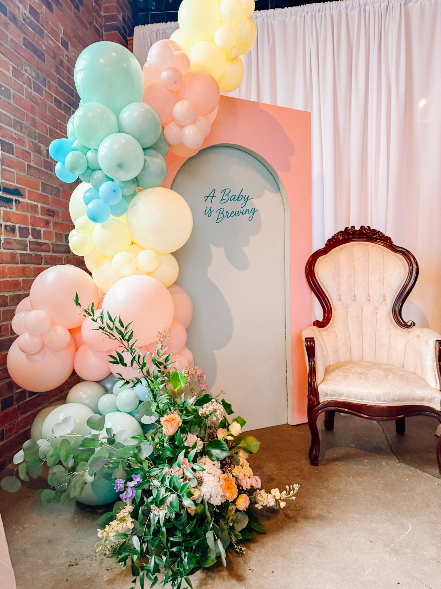 Decorative setup with pastel balloons, a floral arrangement, and a vintage armchair against a pink backdrop with white curtains.