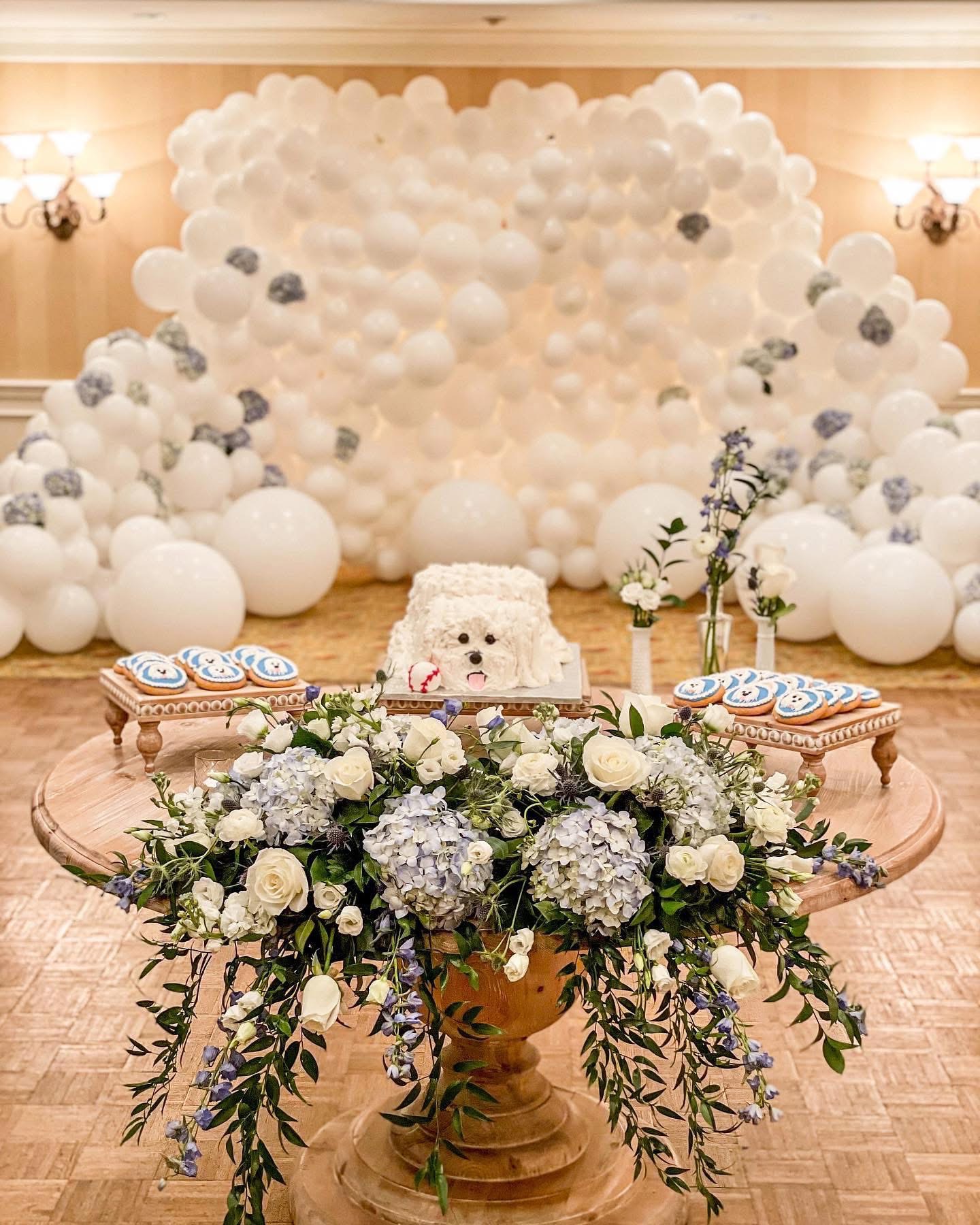 Decorative setup with a large balloon arrangement, a floral centerpiece, and a cake on a table in a decorated room.
