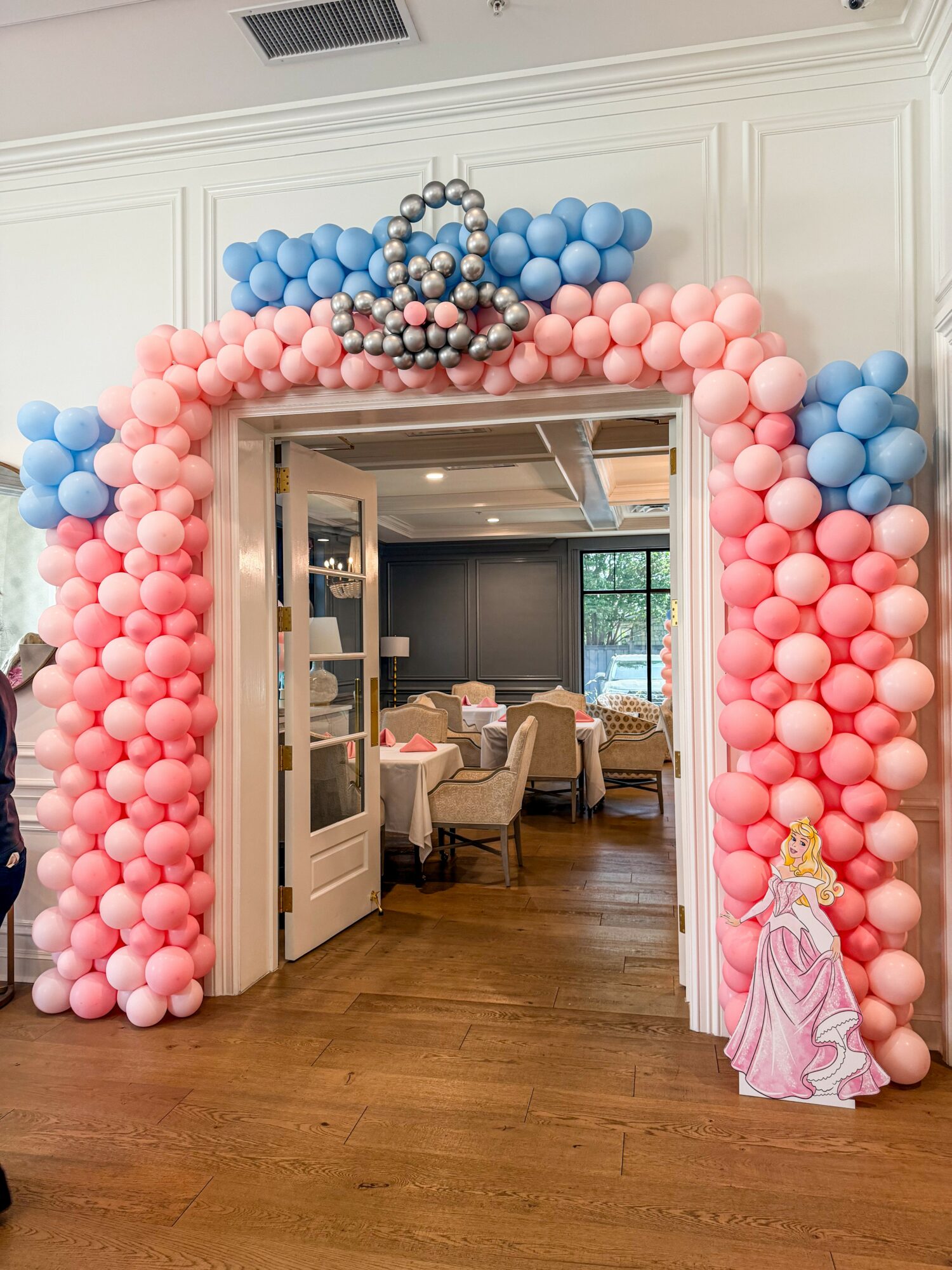 Balloon arch with pink and blue balloons, crown decoration, and a princess figure at the entrance to a dining area.